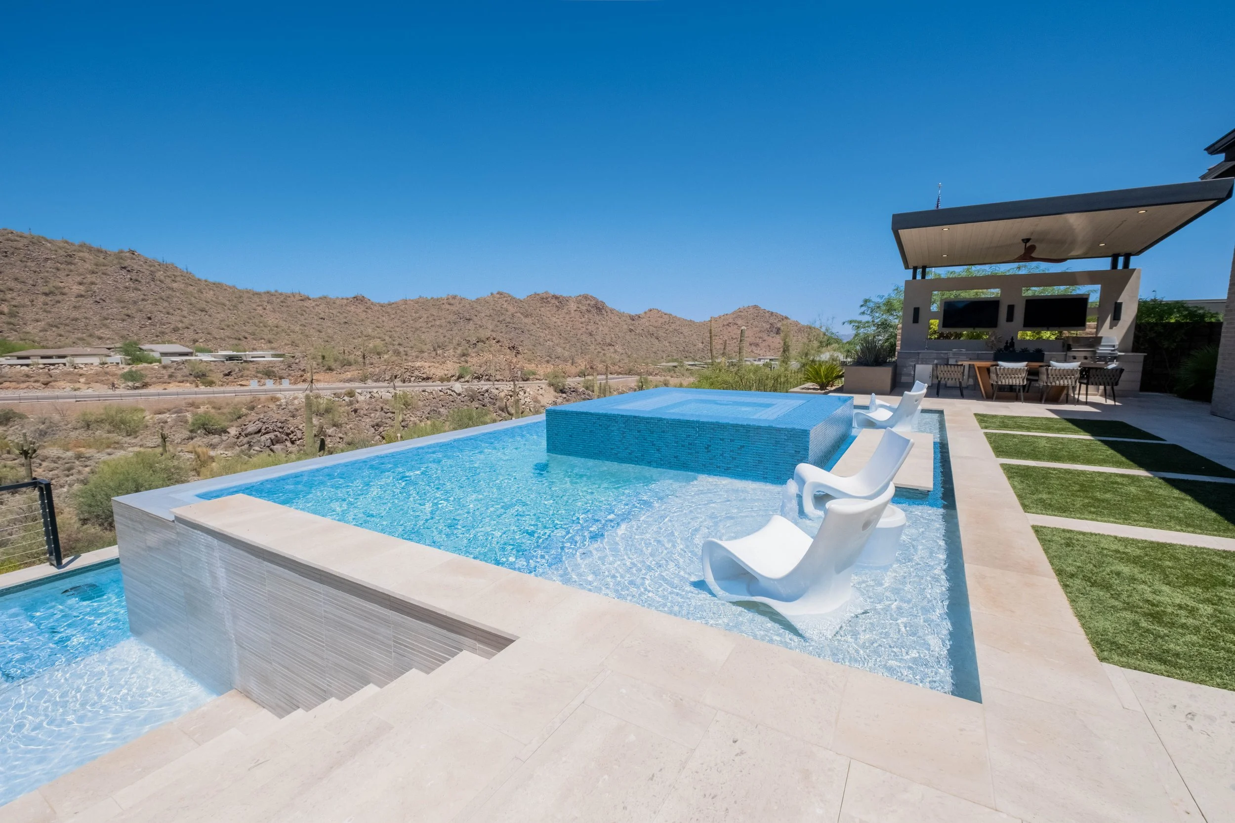 Luxury backyard with a swimming pool and mountain view in the background, featuring poolside chairs, a shaded bar area with multiple TVs, and desert vegetation.