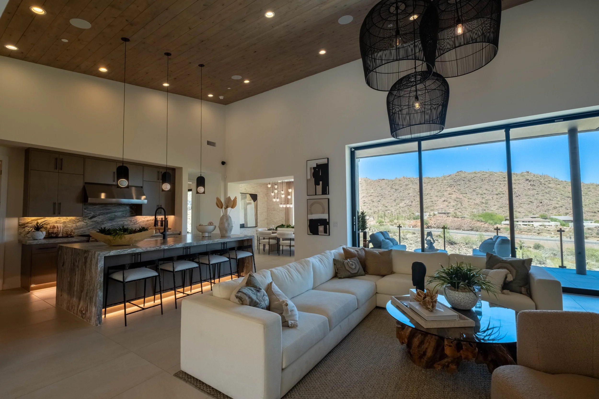 Modern open-concept living room and kitchen with large windows showing desert landscape, featuring a white sectional sofa, black wire pendant lights, wooden ceiling, and island with bar stools.