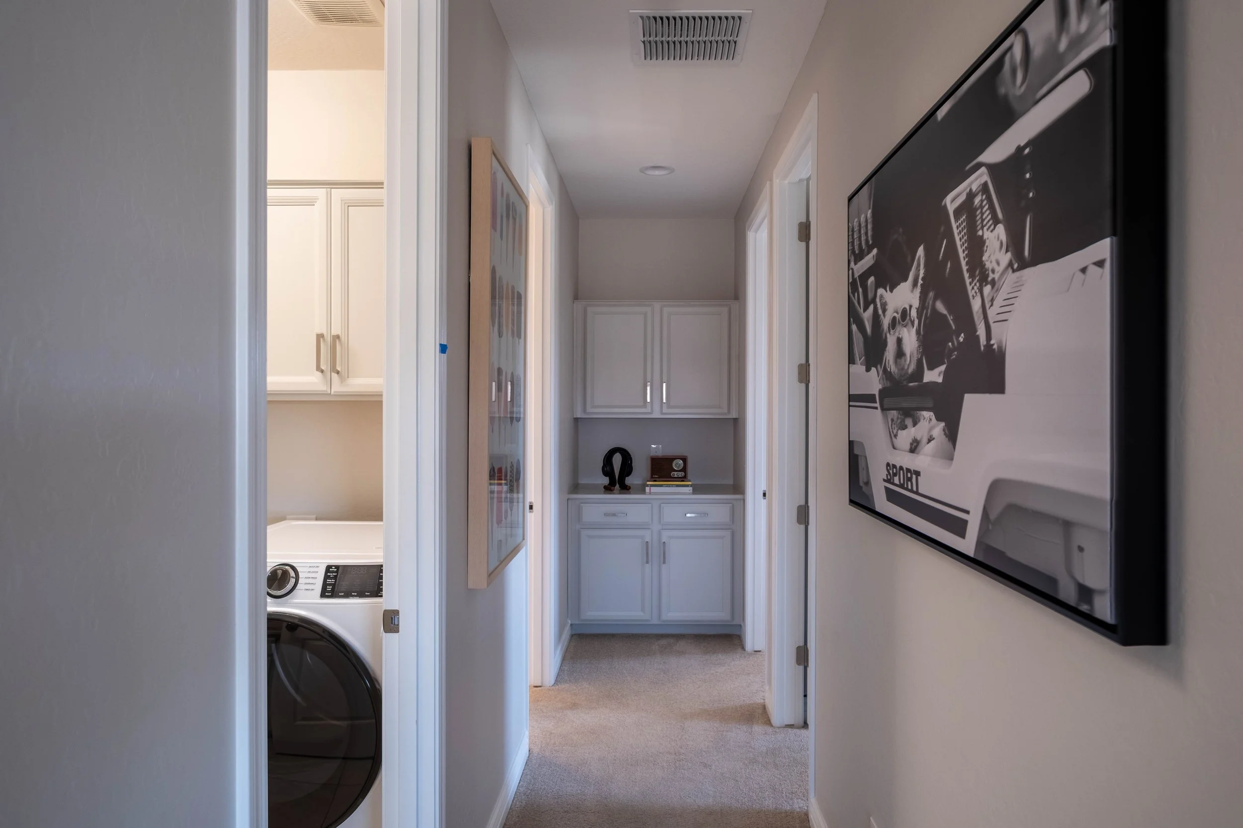 A hallway with white walls and carpeted floor leading to a laundry area and a closet with white cabinets. A washing machine is partially visible on the left, and a black-and-white photograph is mounted on the right wall.