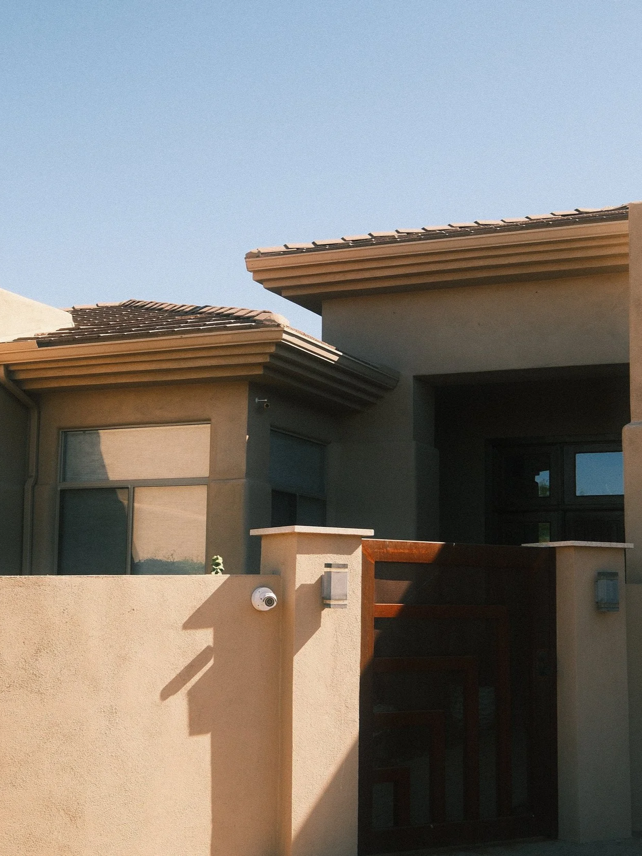 A modern house exterior with beige stucco walls, large windows with shades, decorative ledge, security camera, and wooden door, under a clear blue sky.