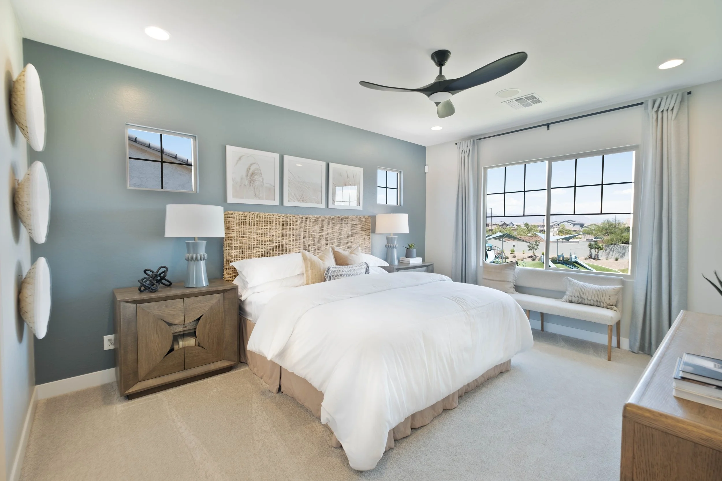 A bedroom with a large bed, nightstands with lamps, framed artwork above the headboard, a window with a view of a suburban neighborhood, and a ceiling fan.