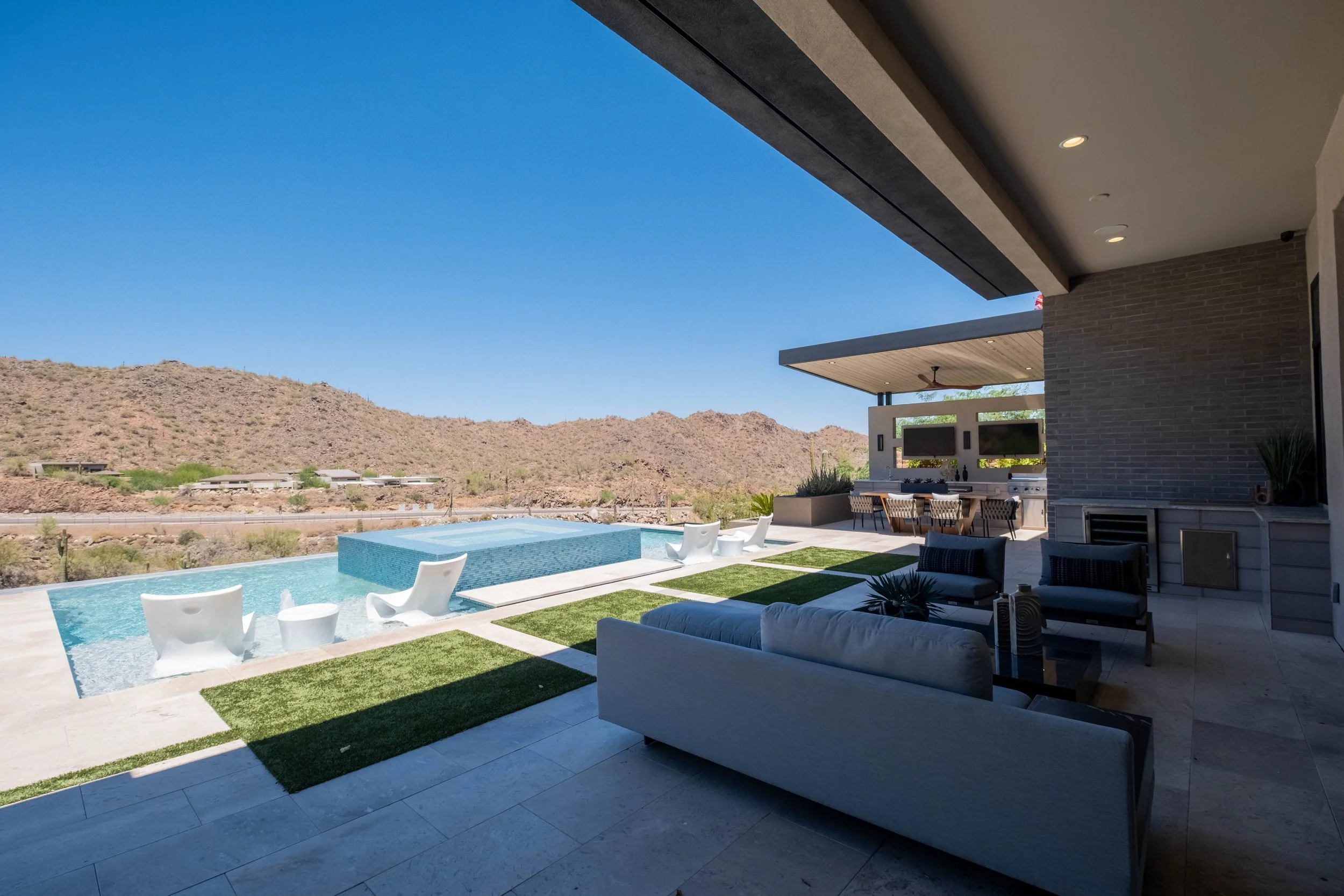 Modern outdoor patio area with a swimming pool, lounge chairs, and a built-in outdoor kitchenset against a backdrop of desert mountains under a clear blue sky.