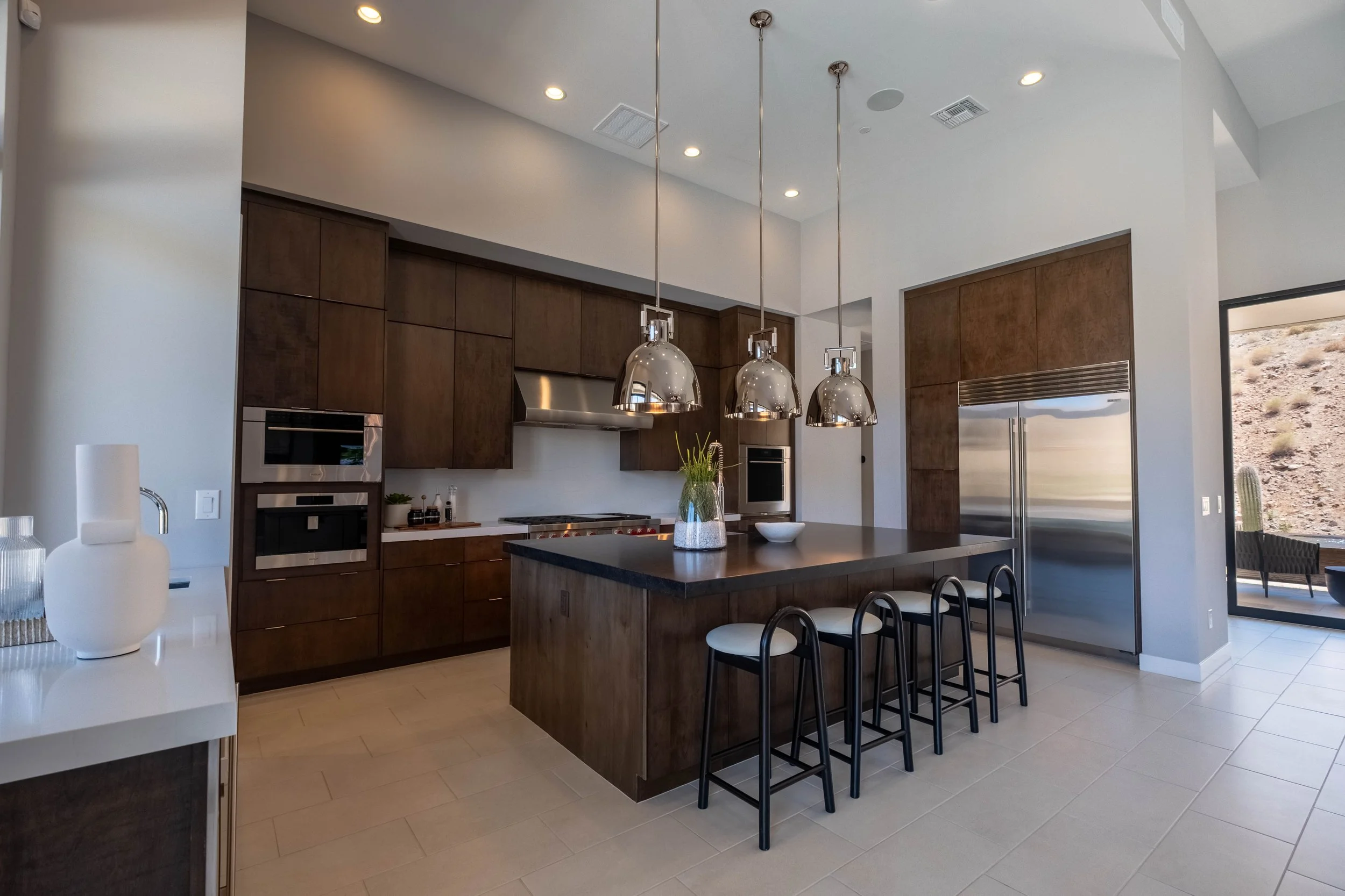Modern kitchen with dark wooden cabinets, a kitchen island with a dark countertop, three hanging metallic pendant lights, stainless steel appliances, and a large window revealing a desert landscape outside.