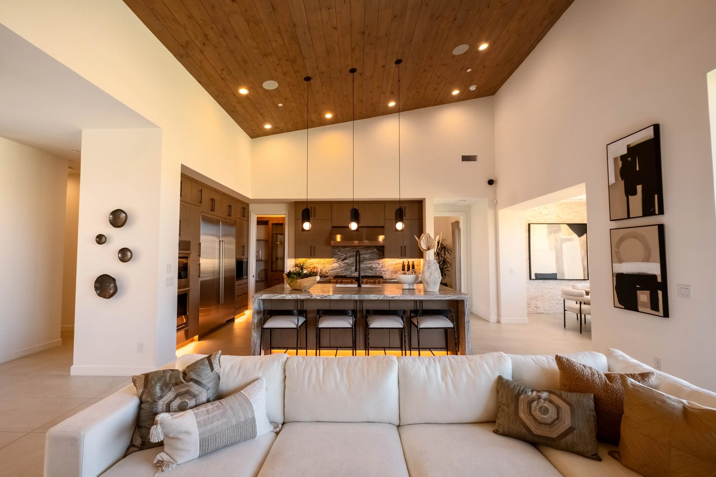 Modern open-concept kitchen and living area with white walls, wooden ceiling, and contemporary decor, featuring a kitchen island with chairs, pendant lighting, and decorative items.
