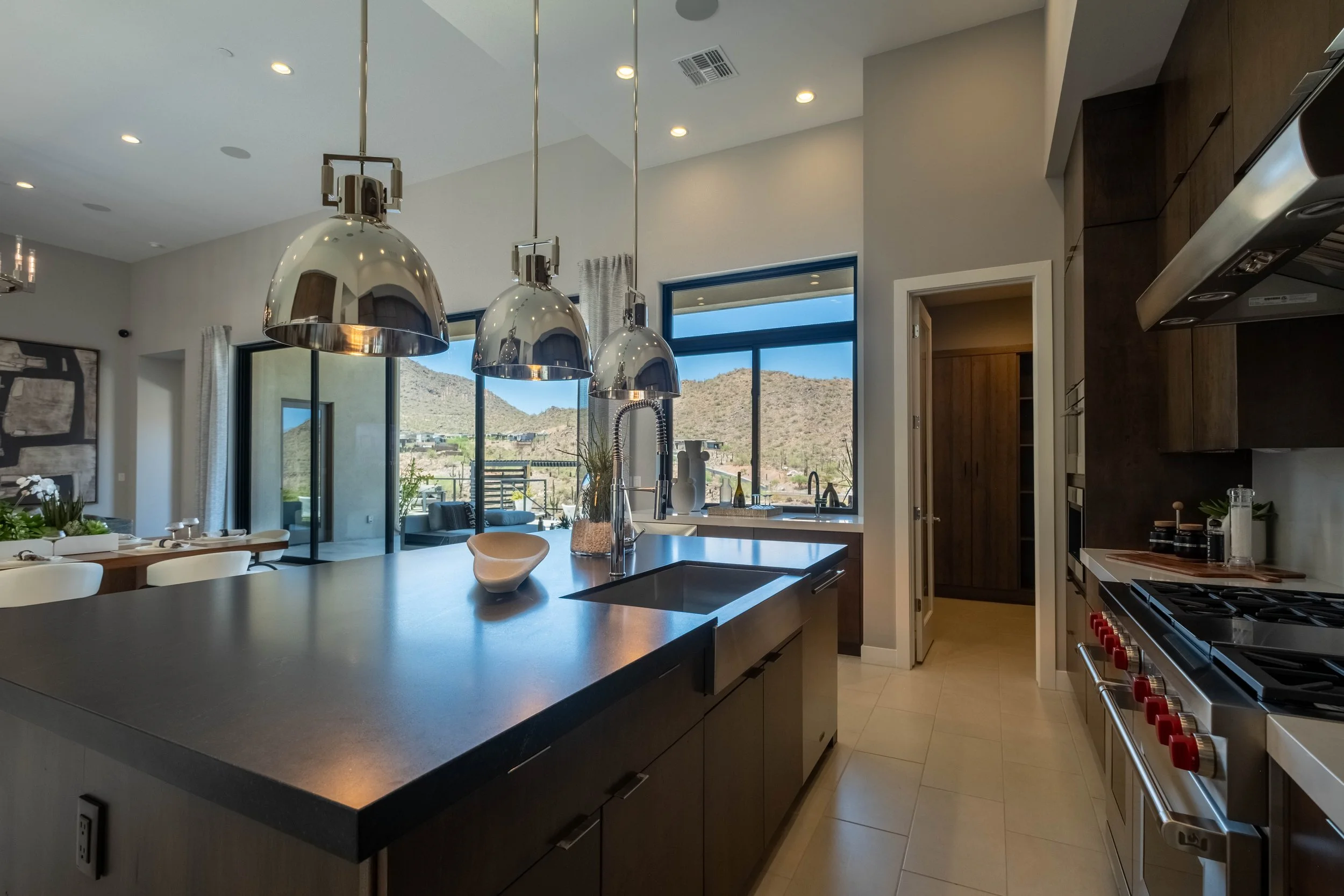 Modern kitchen with large windows revealing mountain view, featuring metallic pendant lights, a dark countertop island, and wooden cabinets.