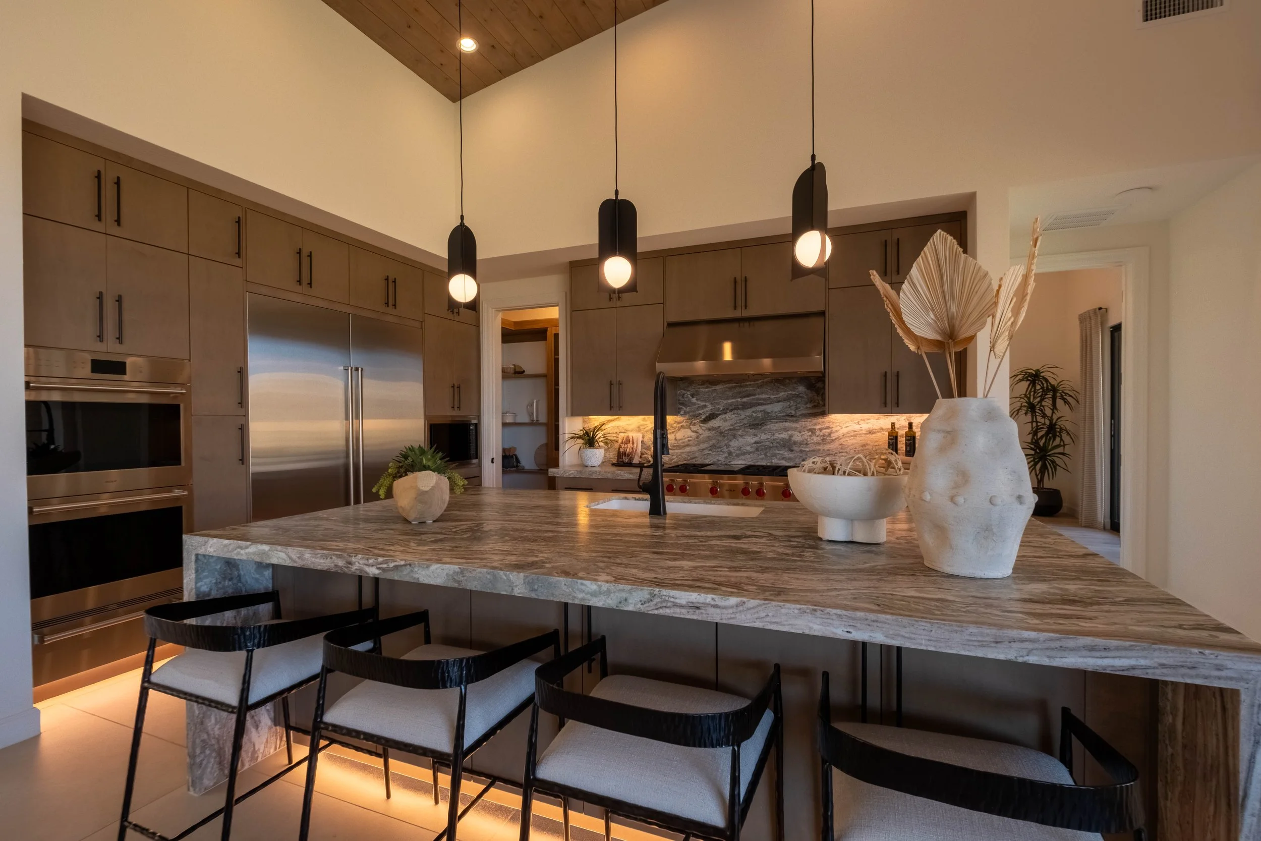 Modern kitchen with wood cabinets, stainless steel refrigerator, marble island, black pendant lights, decorative vases, and dining chairs.