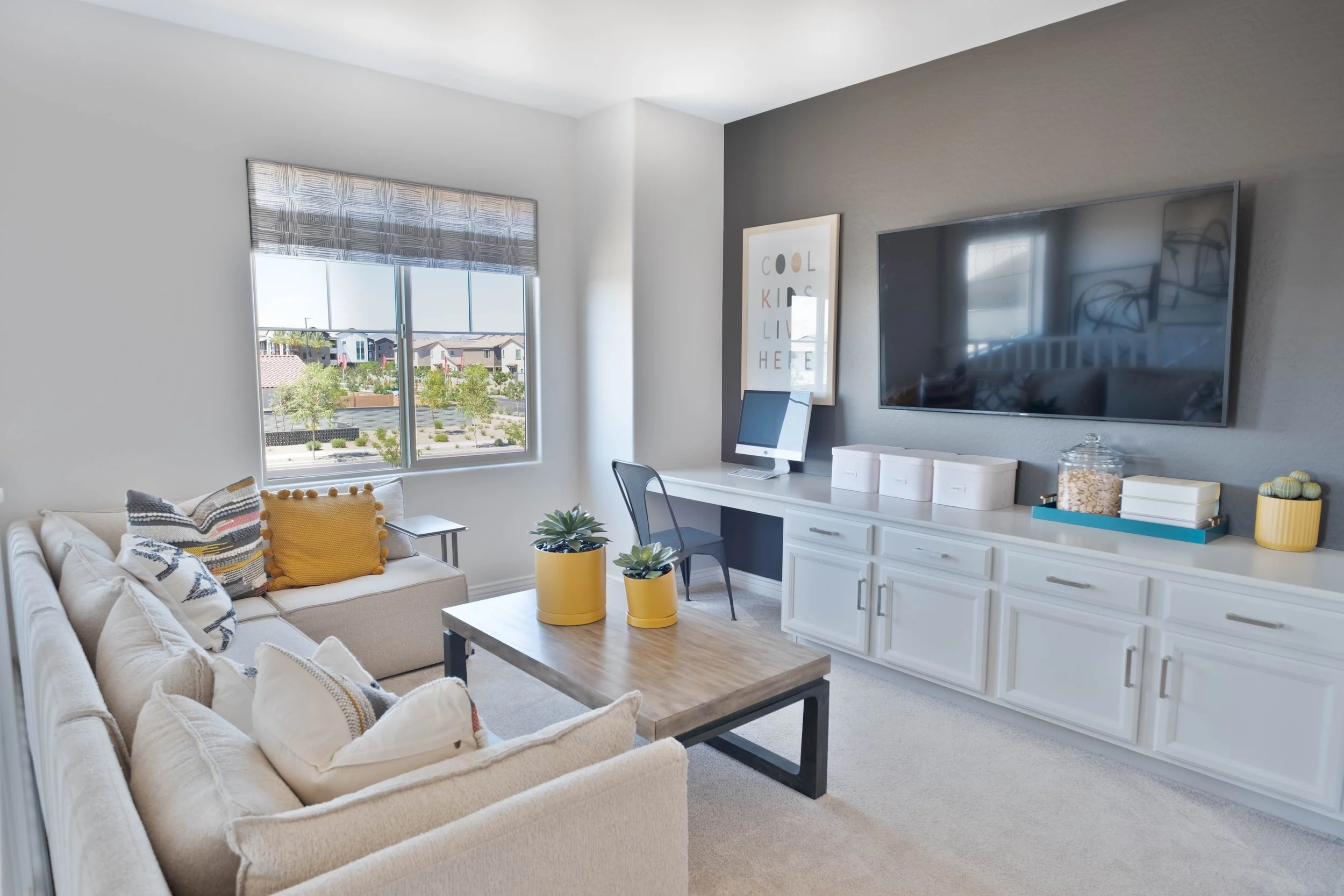 A living room with a beige sofa, patterned pillows, a wooden coffee table, and two yellow potted plants. Large window with a view of houses and trees, mounted flat-screen TV on gray wall, and white cabinet with decorative items.