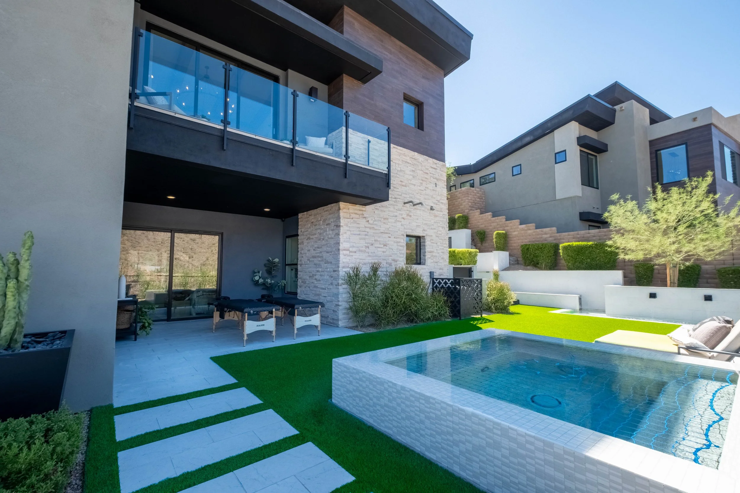 Modern house with a backyard pool area featuring a sitting area, artificial grass, and landscaped plants, with two stories and large glass windows and balconies.