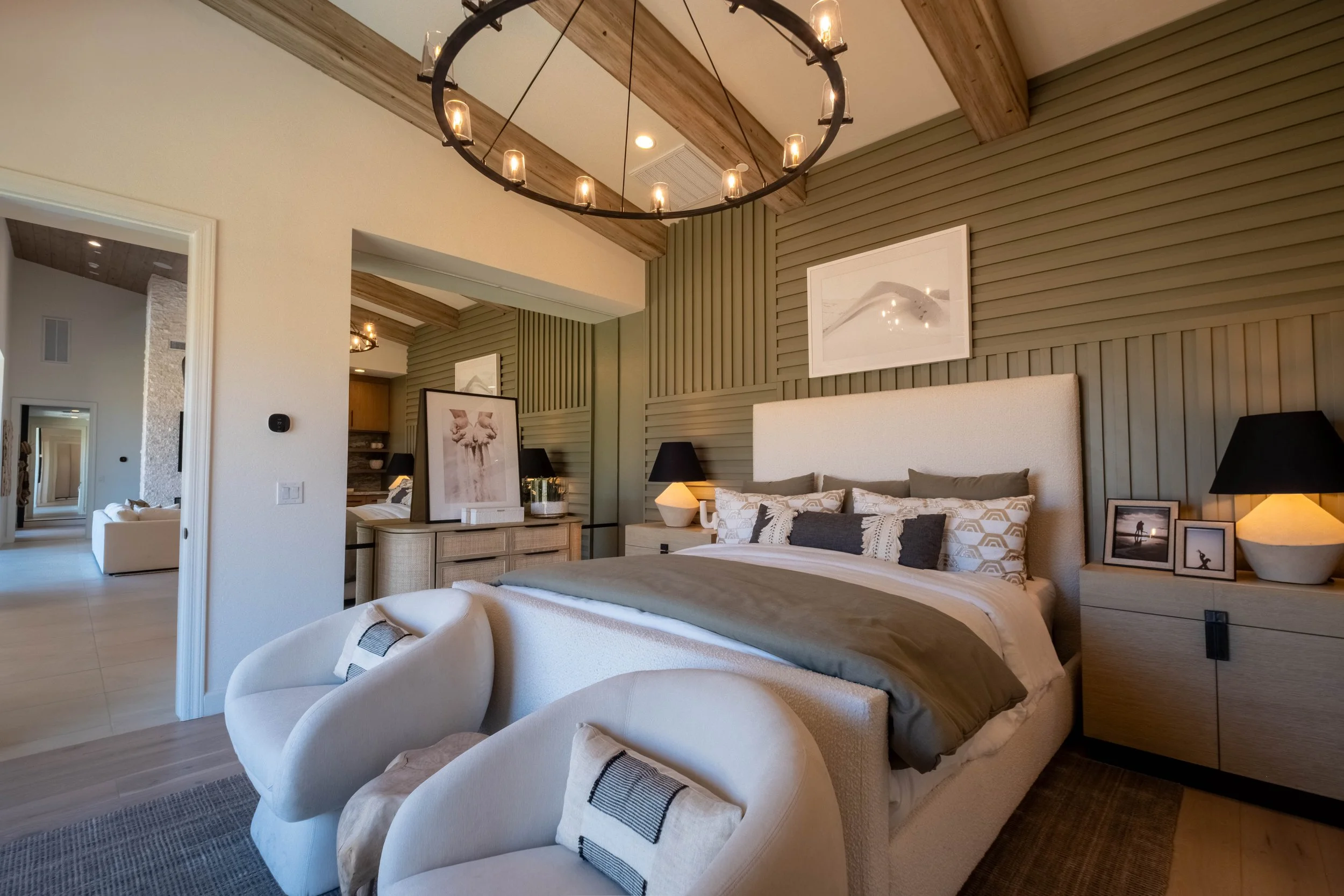 A bedroom with a large bed, a white upholstered headboard, decorative pillows, and a wooden dresser with framed photos and artworks. There's a black table lamp on a nightstand, a modern cream-colored armchair in the foreground, and a chandelier with 