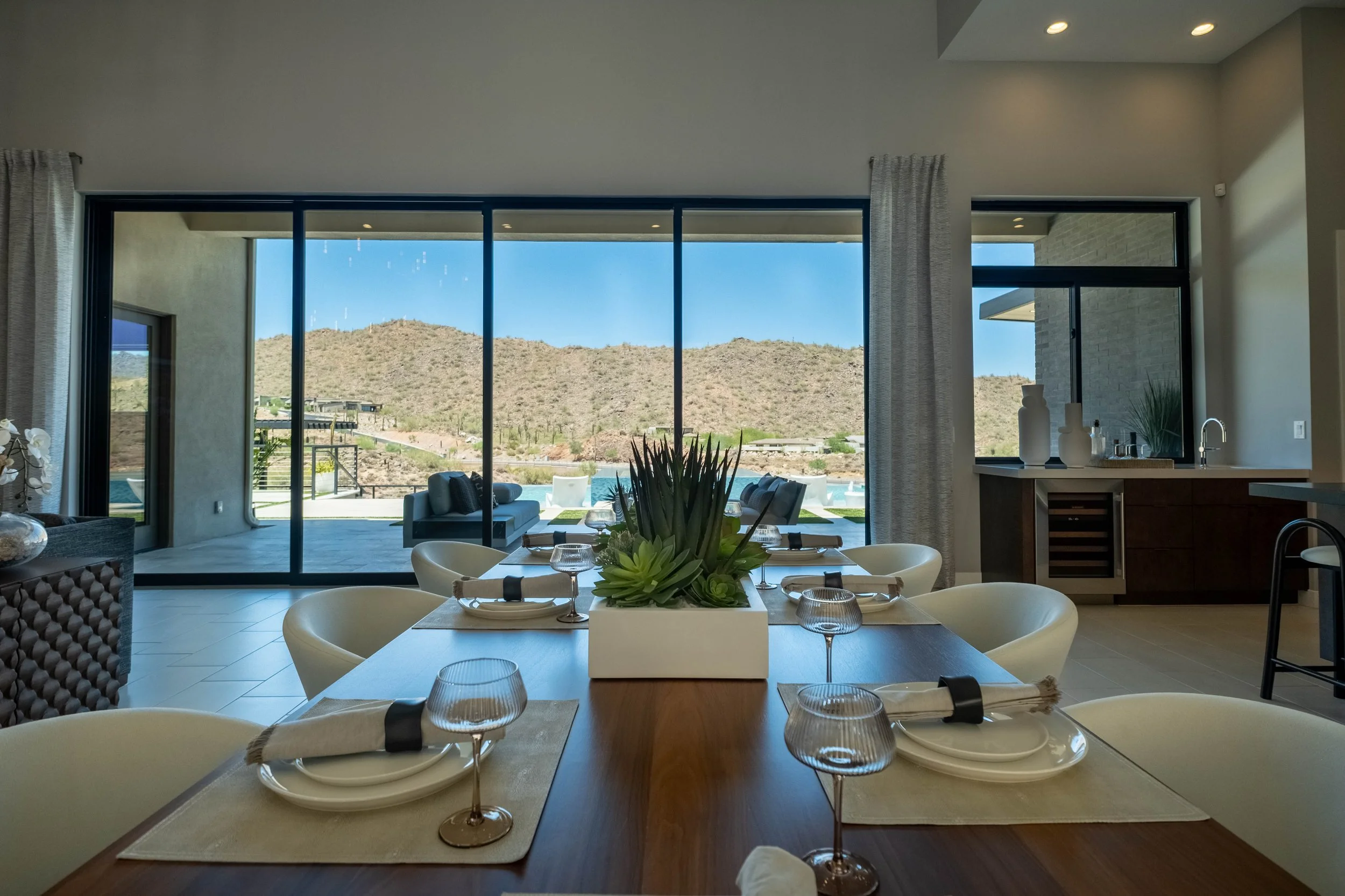 Modern dining room with a large window view of a mountainous landscape, set with a centerpiece of succulents, and surrounded by white chairs and a wooden dining table