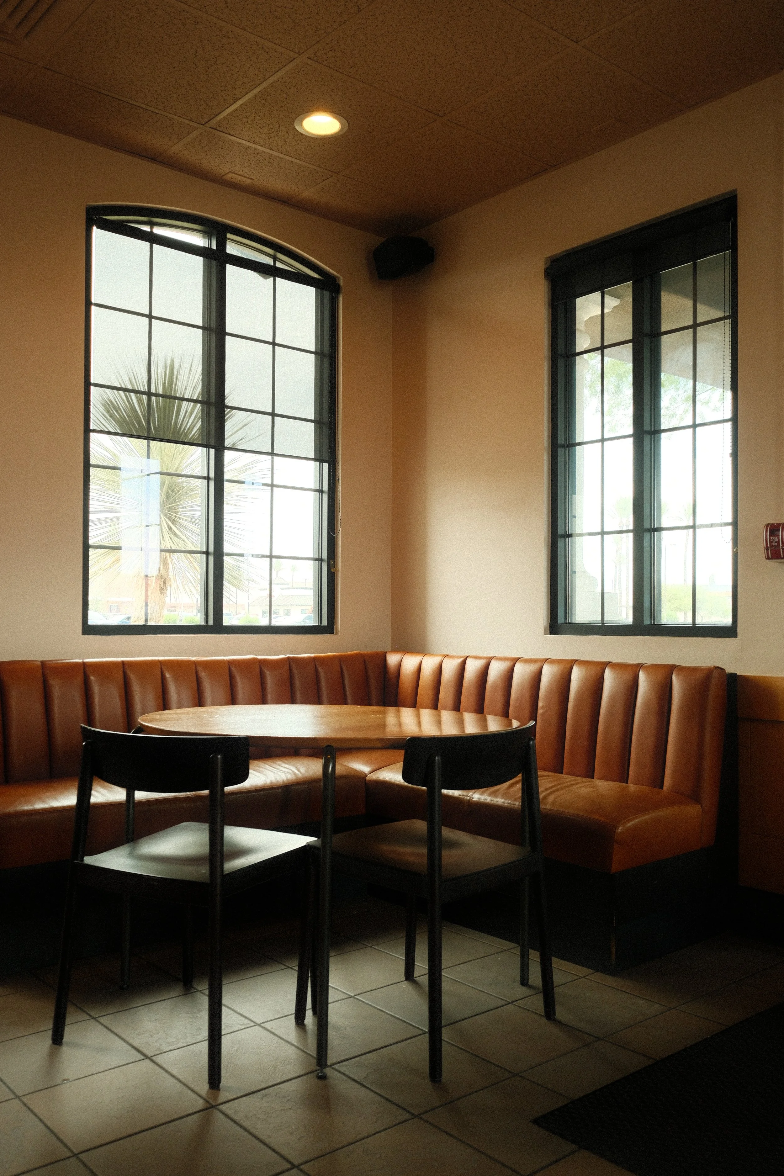Interior of a cafe or restaurant with tile flooring, large windows, a round wooden table, black chairs, and curved tan leather seating.