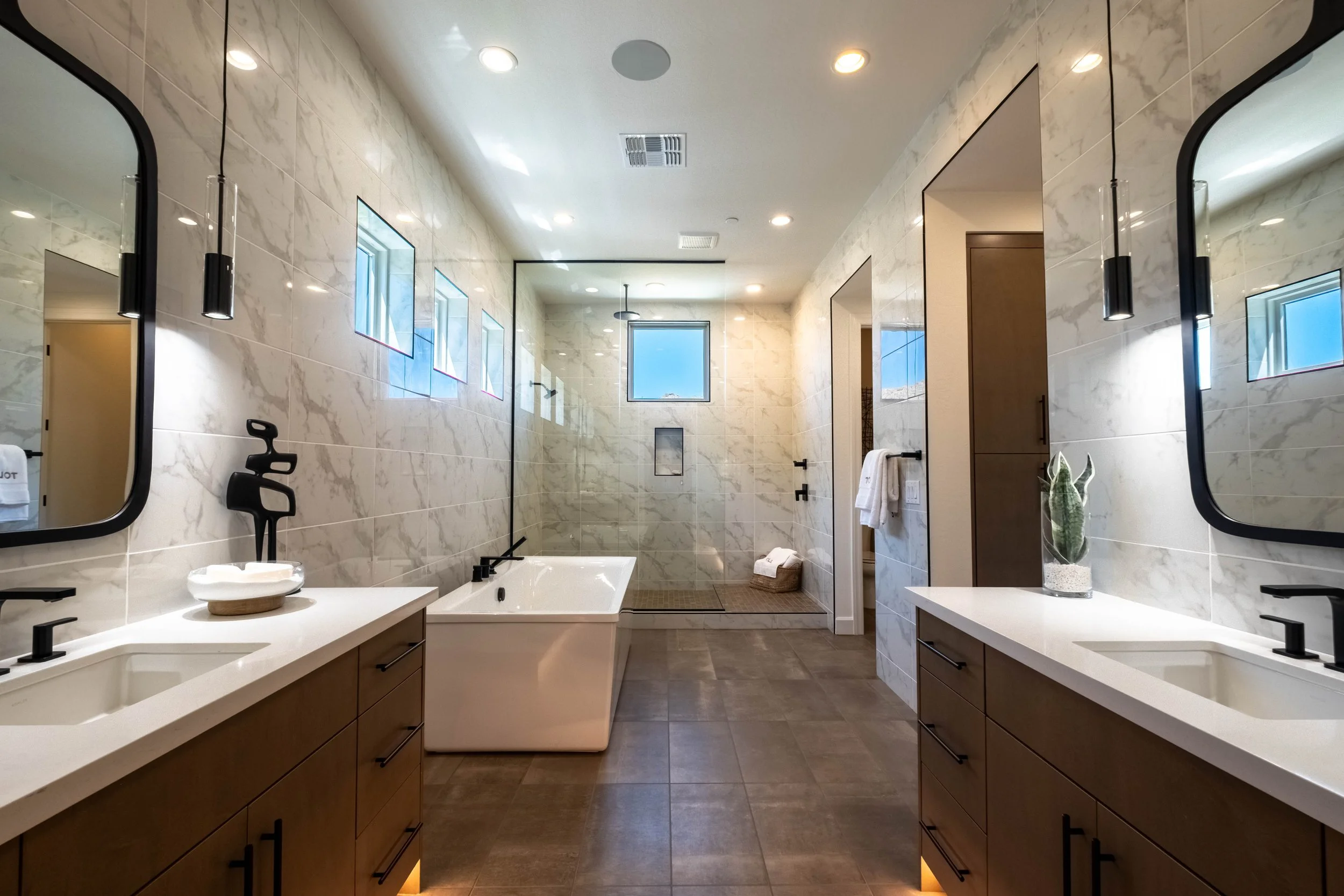 Modern bathroom with marble walls, double sinks with wooden cabinets, large mirrors, pendant lights, a freestanding bathtub, a walk-in shower with glass enclosure, small windows, and a potted plant.