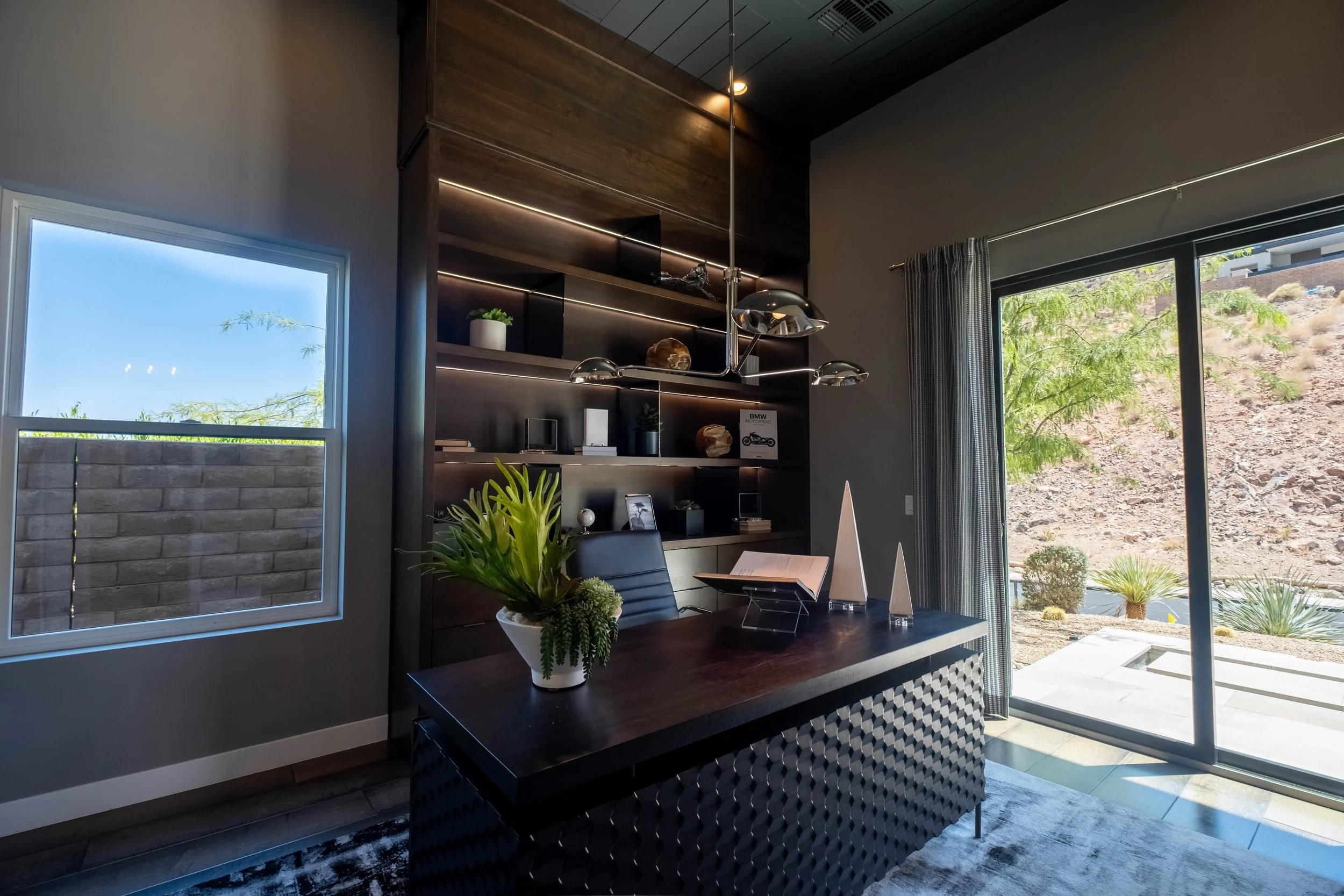 Modern home office with large windows, dark wood shelves, a sleek desk, and a potted plant, overlooking a desert landscape.