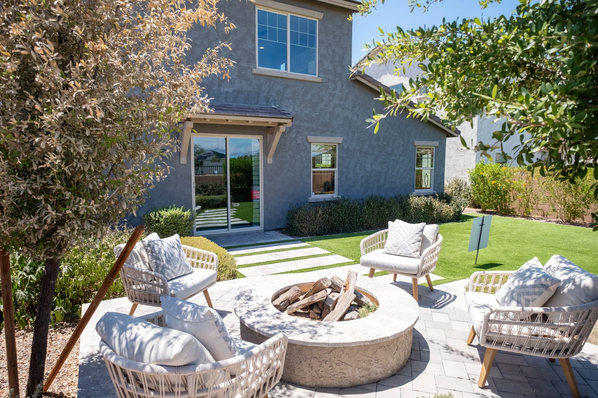 A backyard patio area with white outdoor furniture arranged around a fire pit filled with wood logs. Surrounding the patio are green lawn, shrubs, and trees, with a two-story house in the background featuring large windows and a sliding glass door.