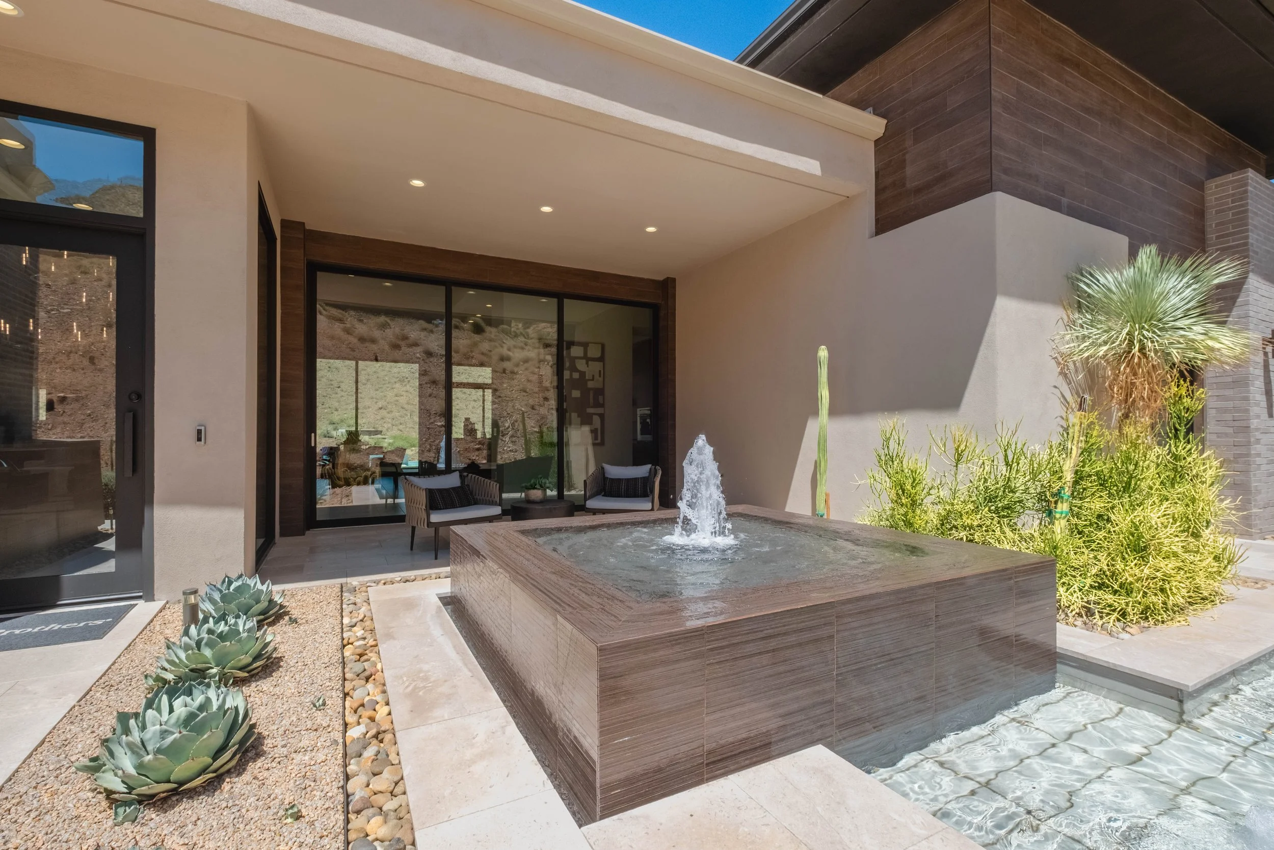 Modern house patio area with a square water fountain, desert plants, and outdoor seating near large glass doors. The house has beige, brown, and dark wood exterior walls.