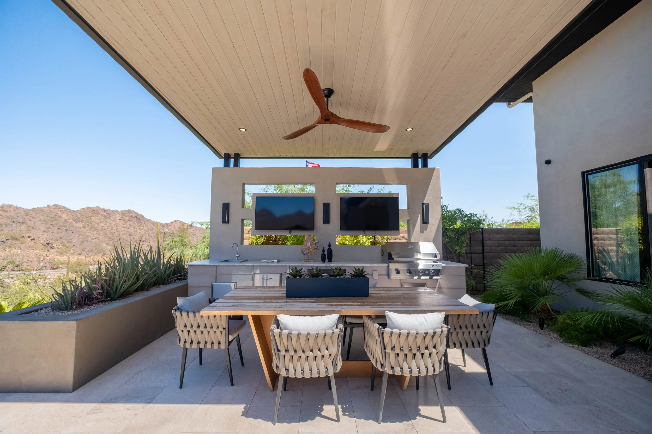 An outdoor patio area with a dining table and chairs, a built-in outdoor kitchen, a ceiling fan, and mountain views in the background.