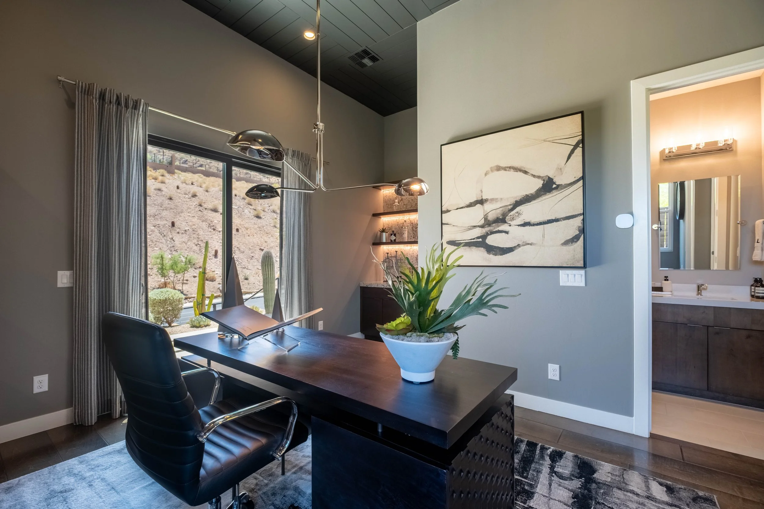 Modern dining room with large window view of desert landscape, dark wood table, black chairs, abstract art on the wall, potted plant centerpiece, and partial view of bathroom mirror and lighting.