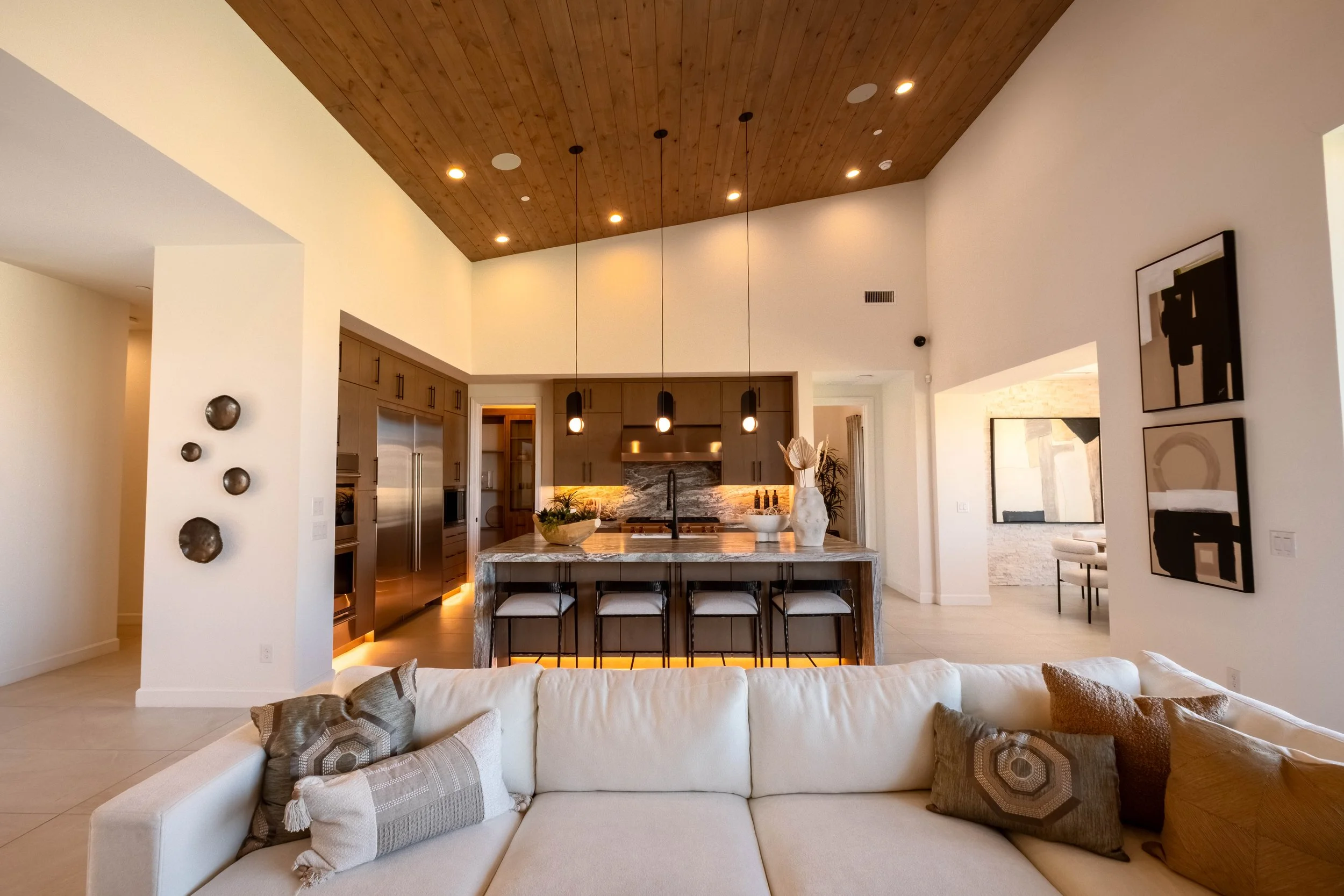 A modern open-concept living space with a kitchen island, stools, and a clear view into the kitchen area with wood cabinets and stainless steel appliances. There's a ceiling with recessed lighting and hanging pendant lights, and some artwork on the wall.