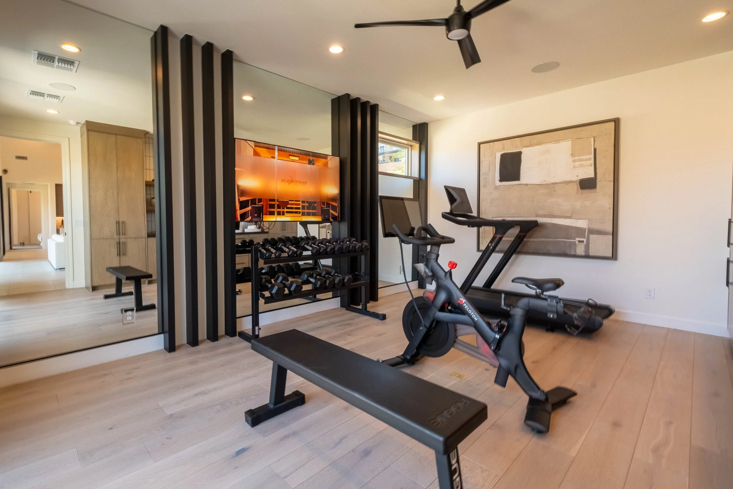 Home gym with a treadmill, exercise bike, dumbbell rack, mirror, artwork on white walls, and black ceiling fan.