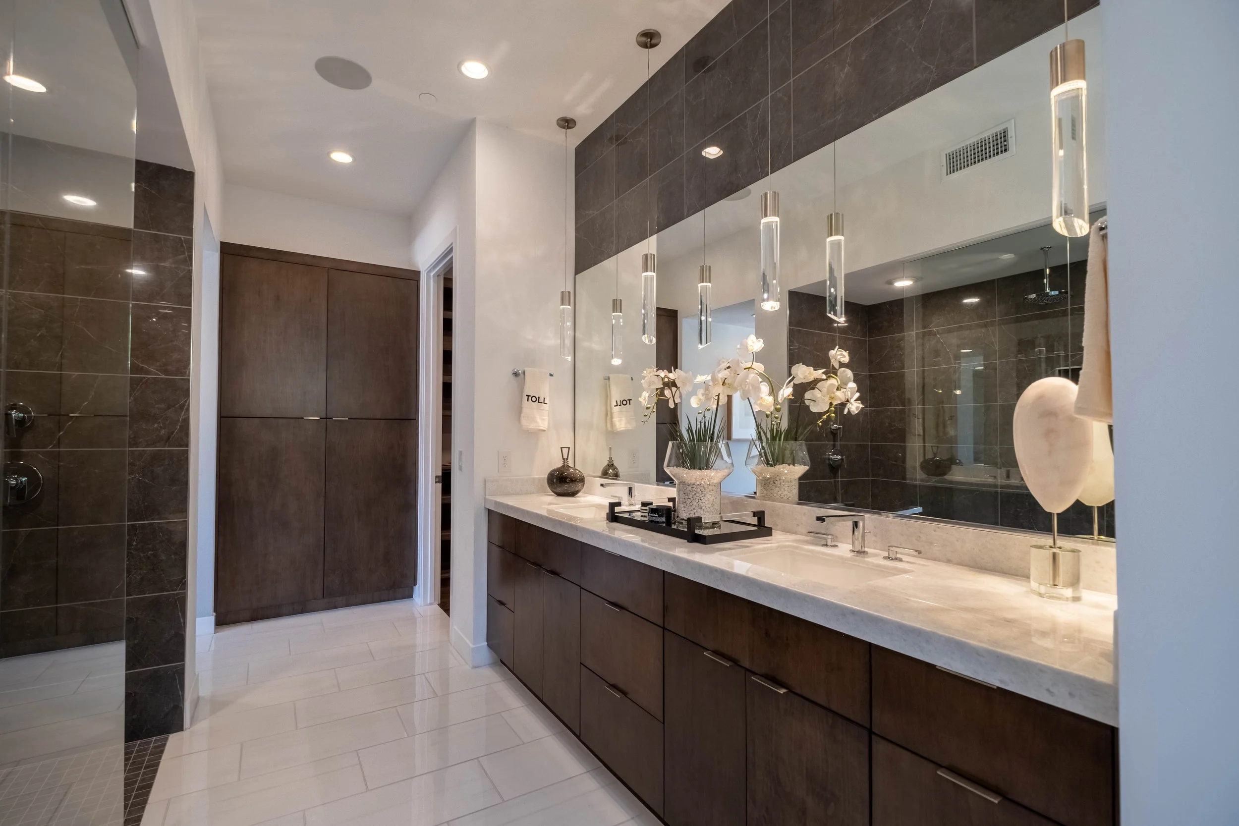 Modern bathroom with dark wood vanity, large mirror, white countertop, and decorative plants. Recessed lighting and pendant lights hang above the mirror.