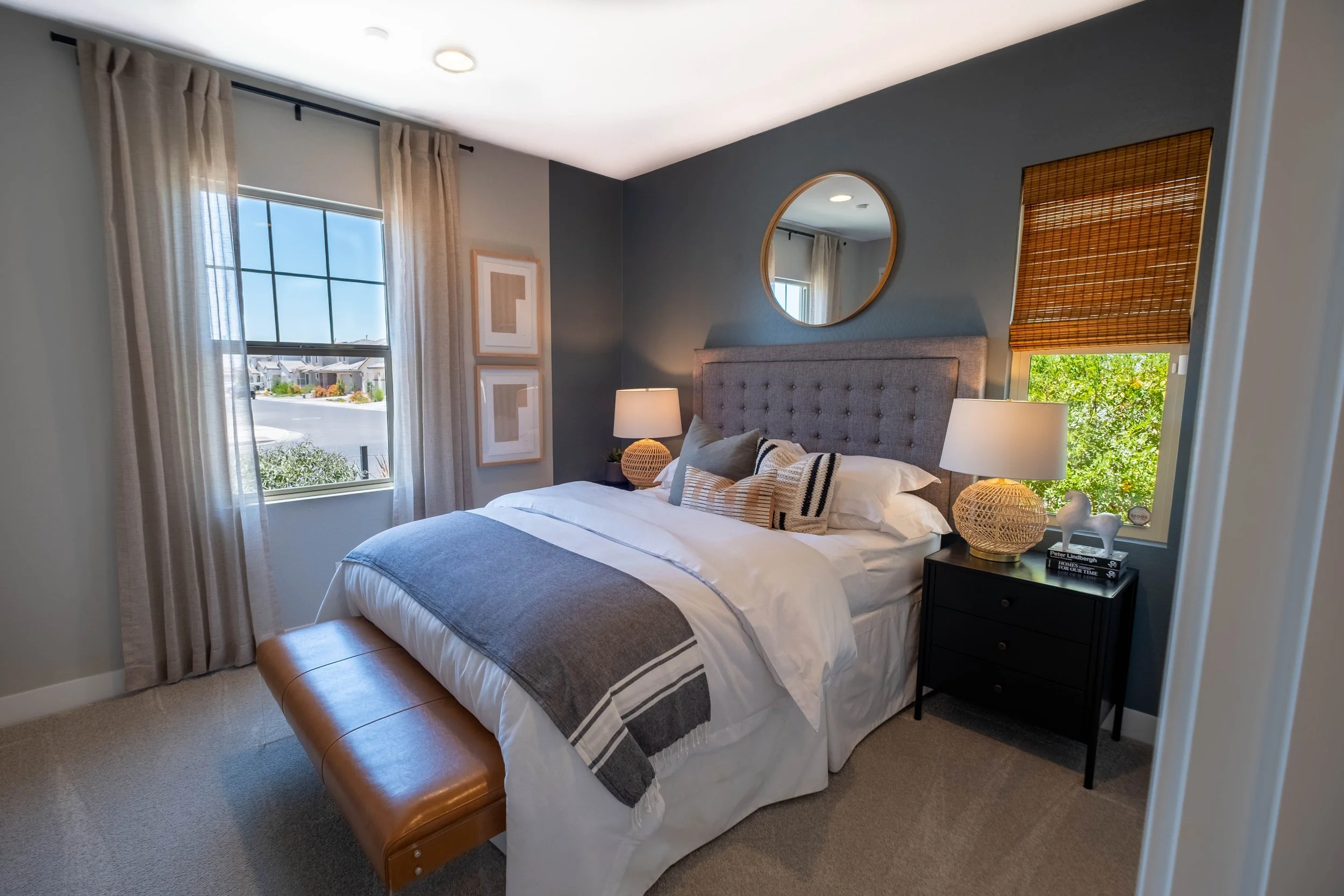 A bedroom with a bed, nightstand, and window with beige curtains, blue walls, and a mirror above the bed.