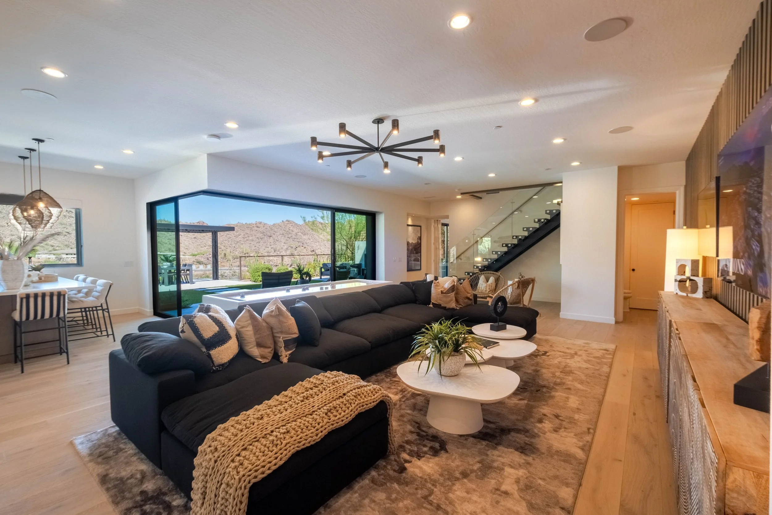 Modern living room with large sectional sofa, coffee tables, indoor plants, and large glass doors leading to an outdoor patio with mountain view.