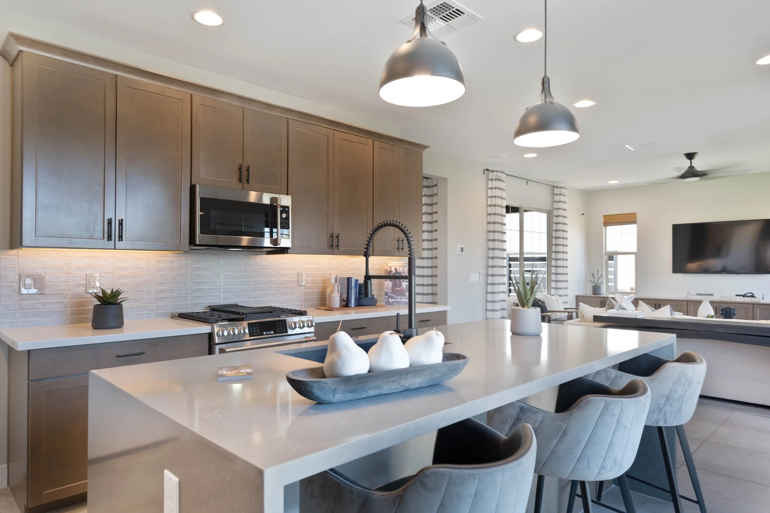 Modern kitchen with wood cabinets, stainless steel microwave and oven, white countertops, black faucet, and open living area with sofa and TV.