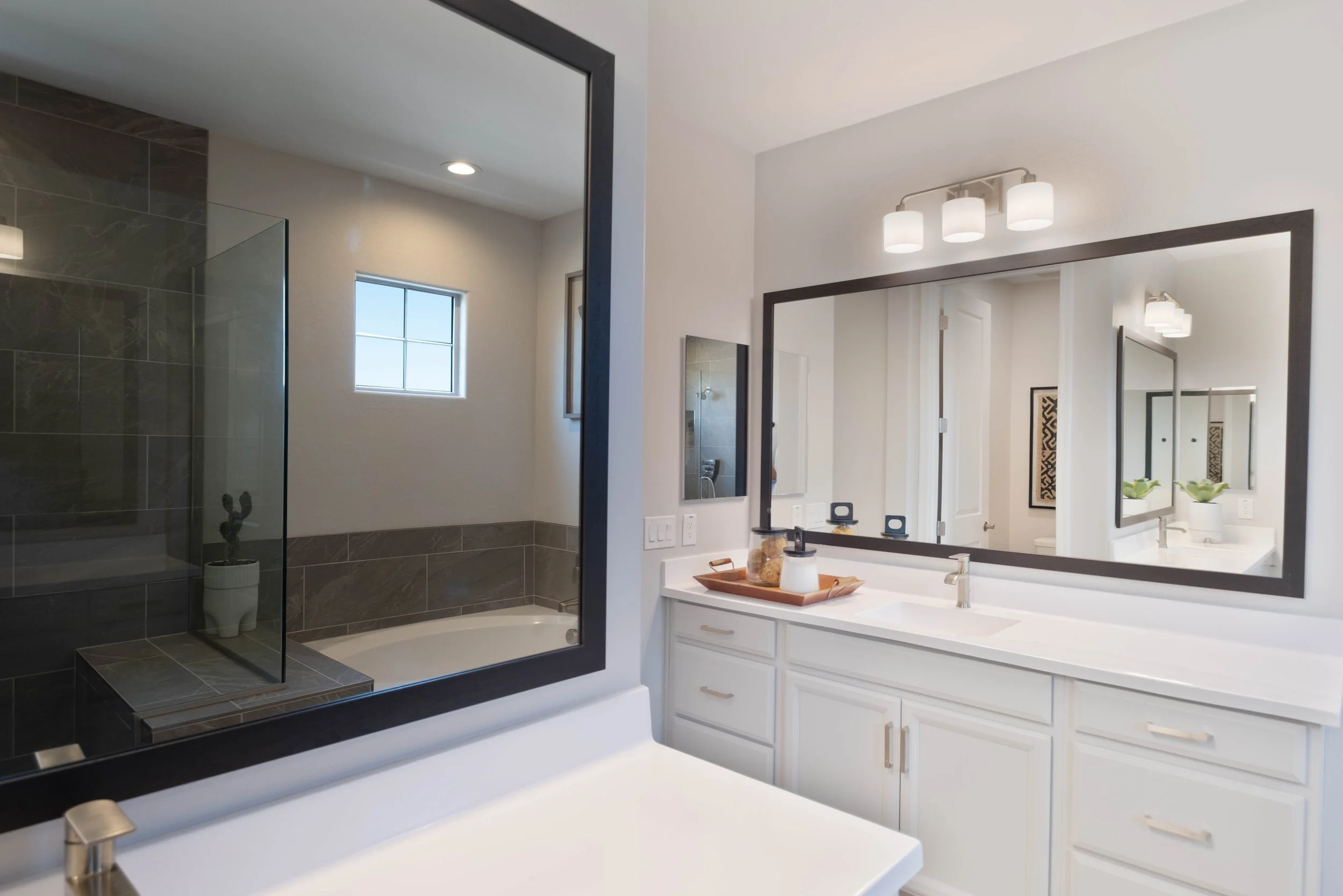A bathroom with a large white vanity, a big mirror, and a modern light fixture with three white shades above it. There are small decorative items on the counter. To the left, there is a walk-in shower with dark grey and brown tiles and a glass door. 