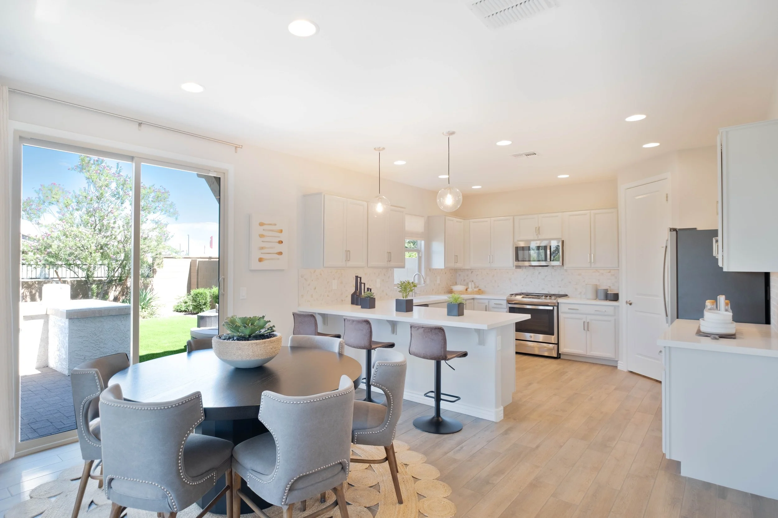 Bright, modern kitchen with white cabinets, pendant lighting, stainless steel appliances, a marble island, and dining table with upholstered chairs, large sliding glass door showing backyard with grass and trees.