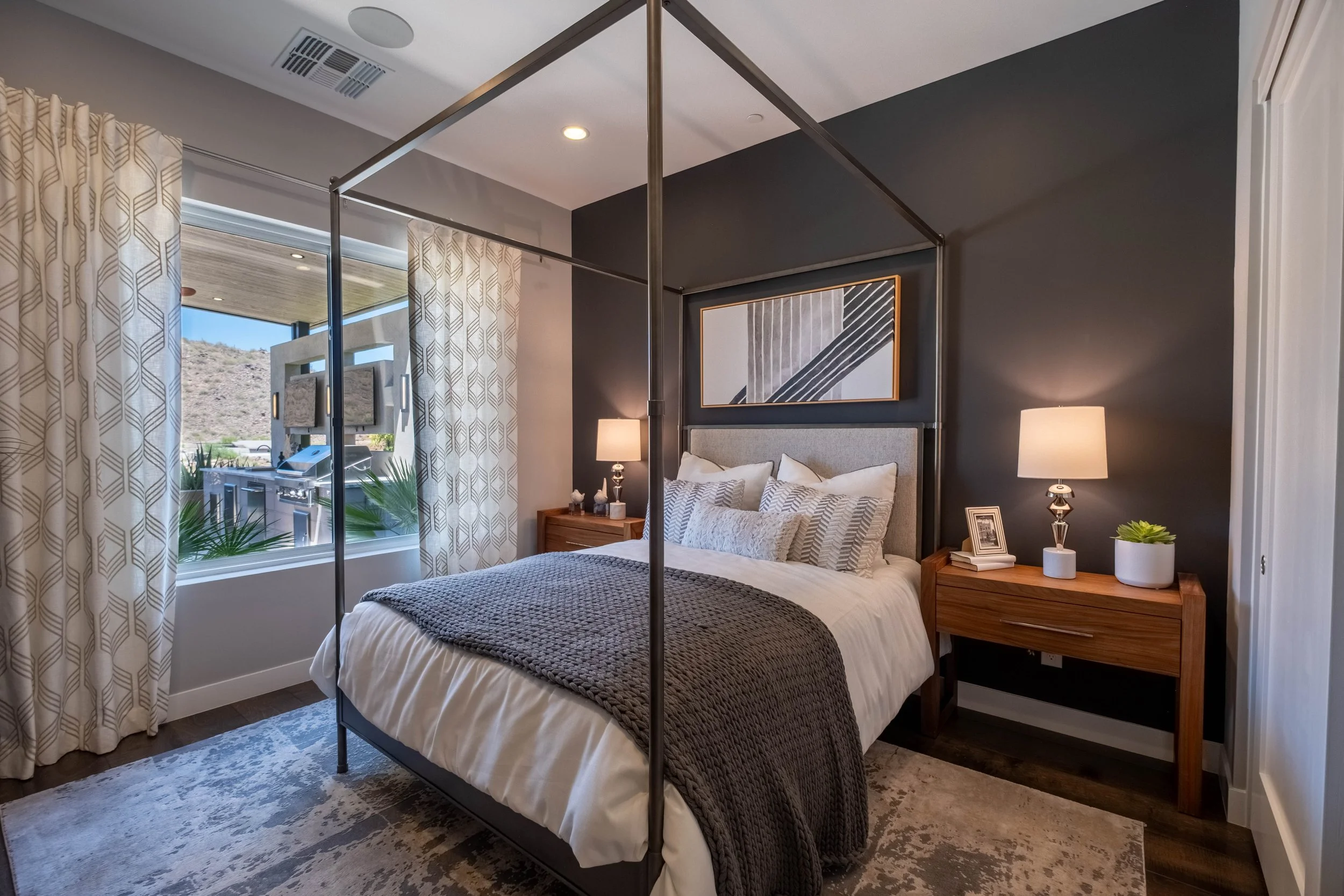 A bedroom with a four-poster bed, gray bedding, and decorative pillows. Two wooden nightstands with lamps and decor. Large window with patterned curtains and a framed modern art piece above the bed.