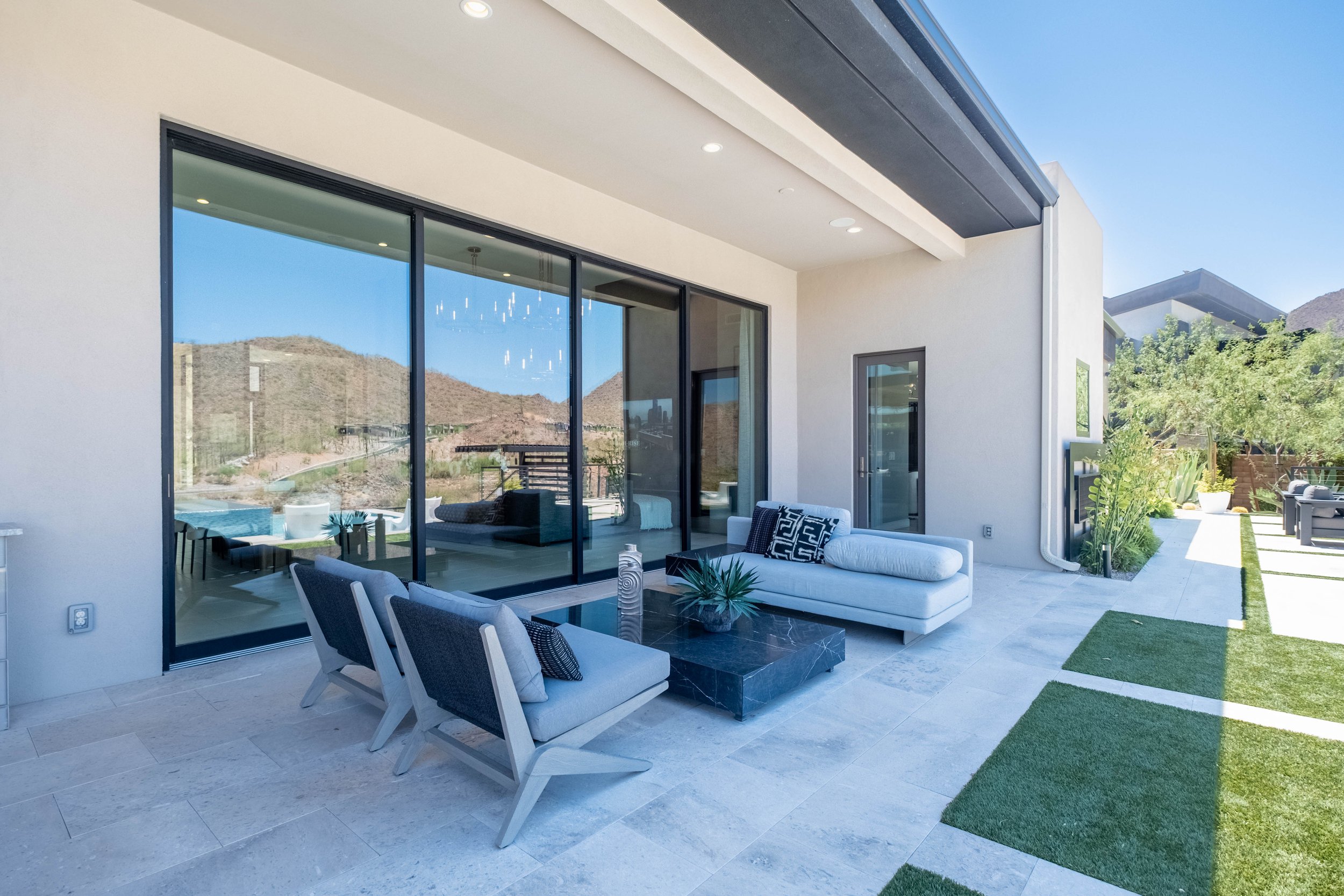 Modern outdoor patio with seating, dining table, large glass doors, and a view of hills in the background, featuring a white sofa, armchair, and green plants.