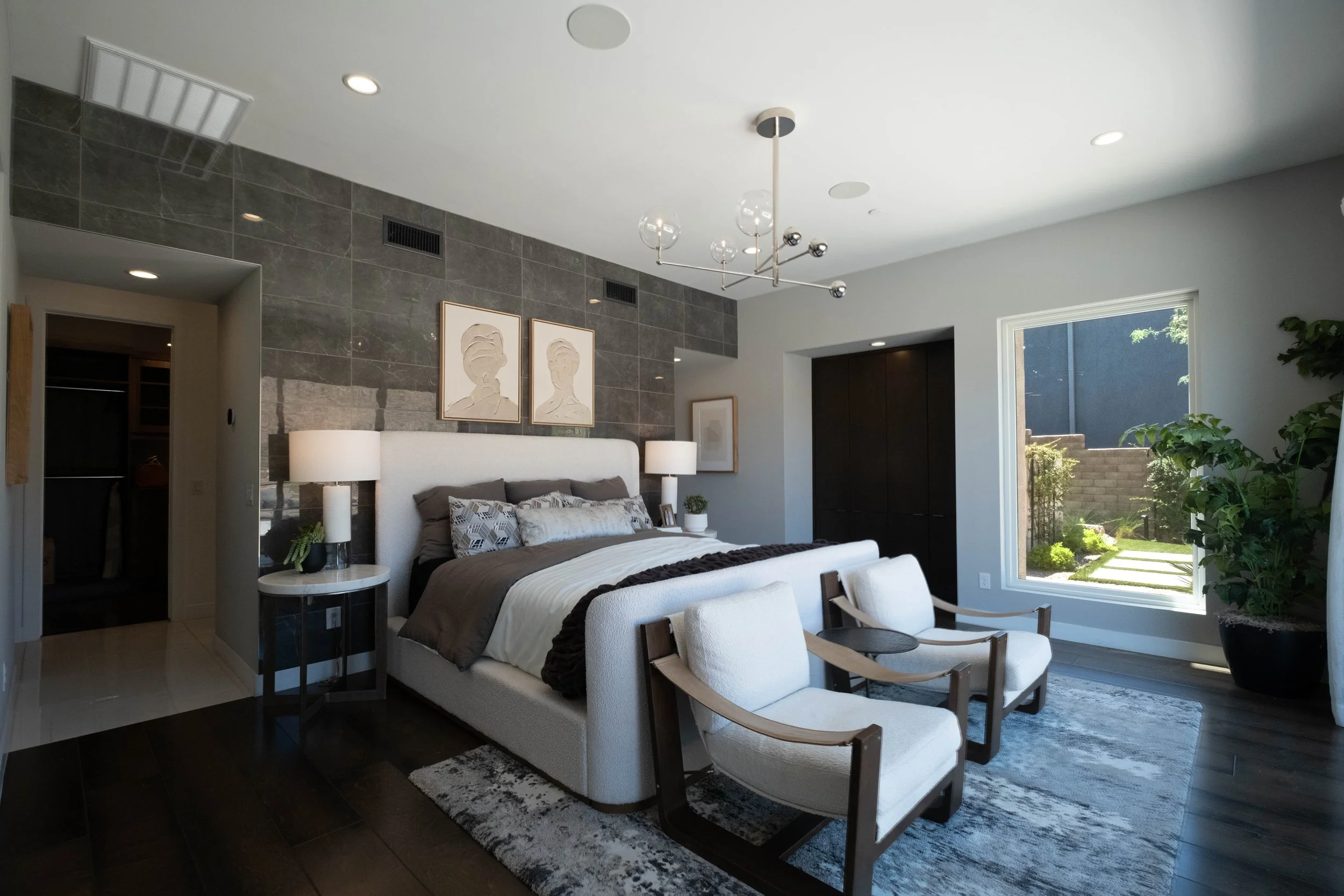 Modern bedroom with a large bed, white headboard, bedside lamps, framed artwork, a sitting chair, window with a view of a garden, and a chandelier with glass globes.