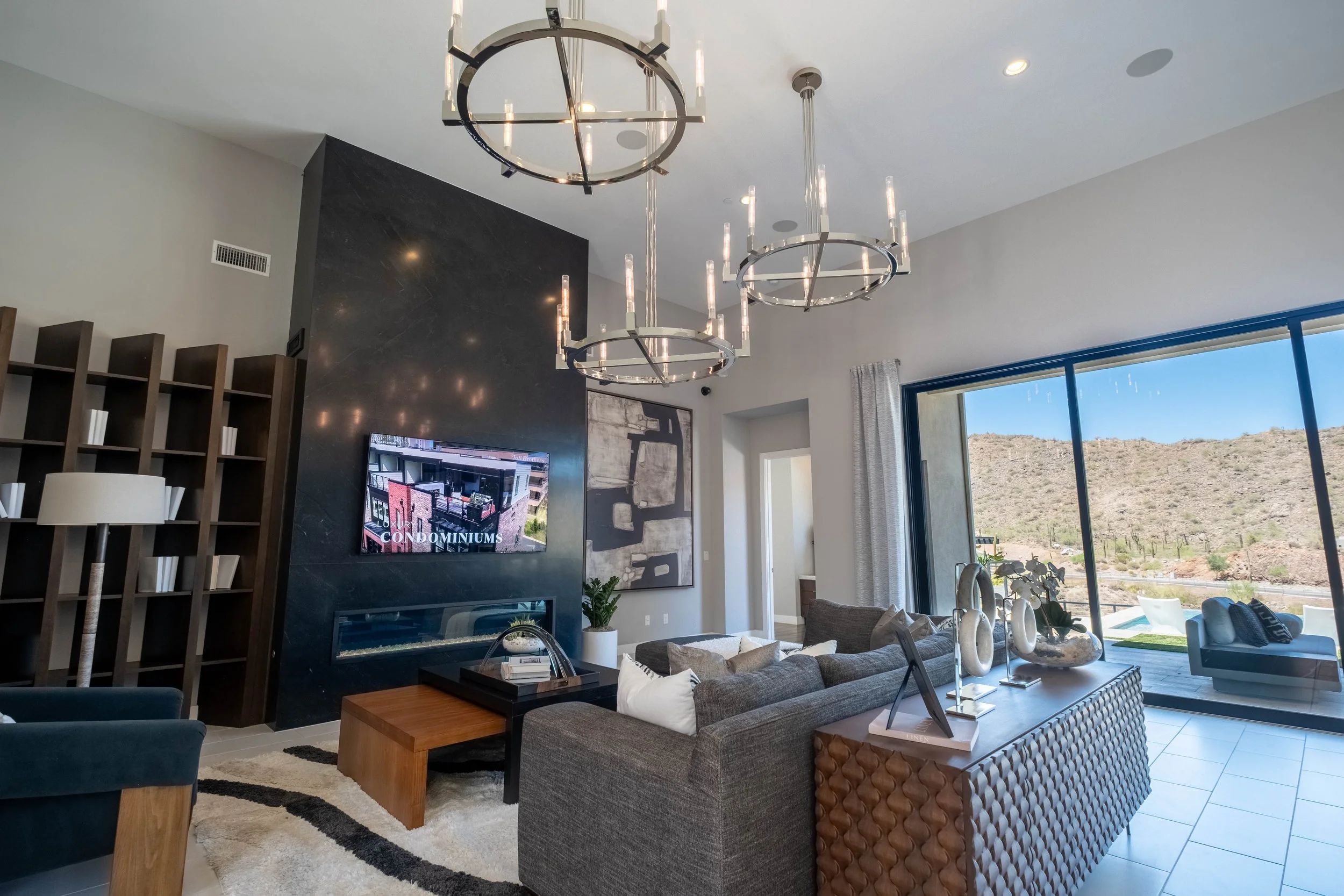 Modern living room with a black feature wall, a flat-screen TV, and contemporary light fixtures hanging from the ceiling. Large windows reveal a desert landscape outside.