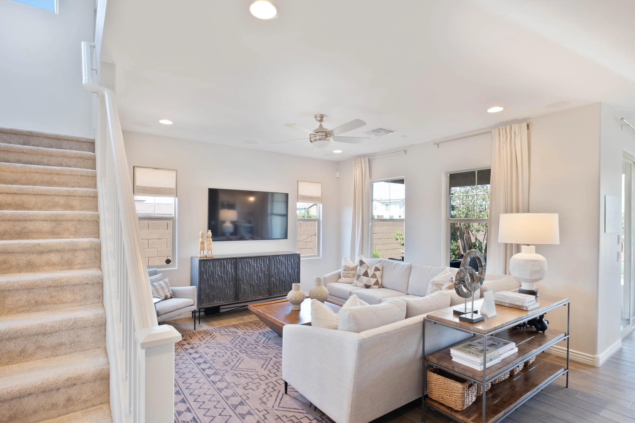 Bright living room with a beige sectional sofa, a coffee table, a large flat-screen TV on the wall, and windows with light curtains, seen from the bottom of a carpeted staircase.
