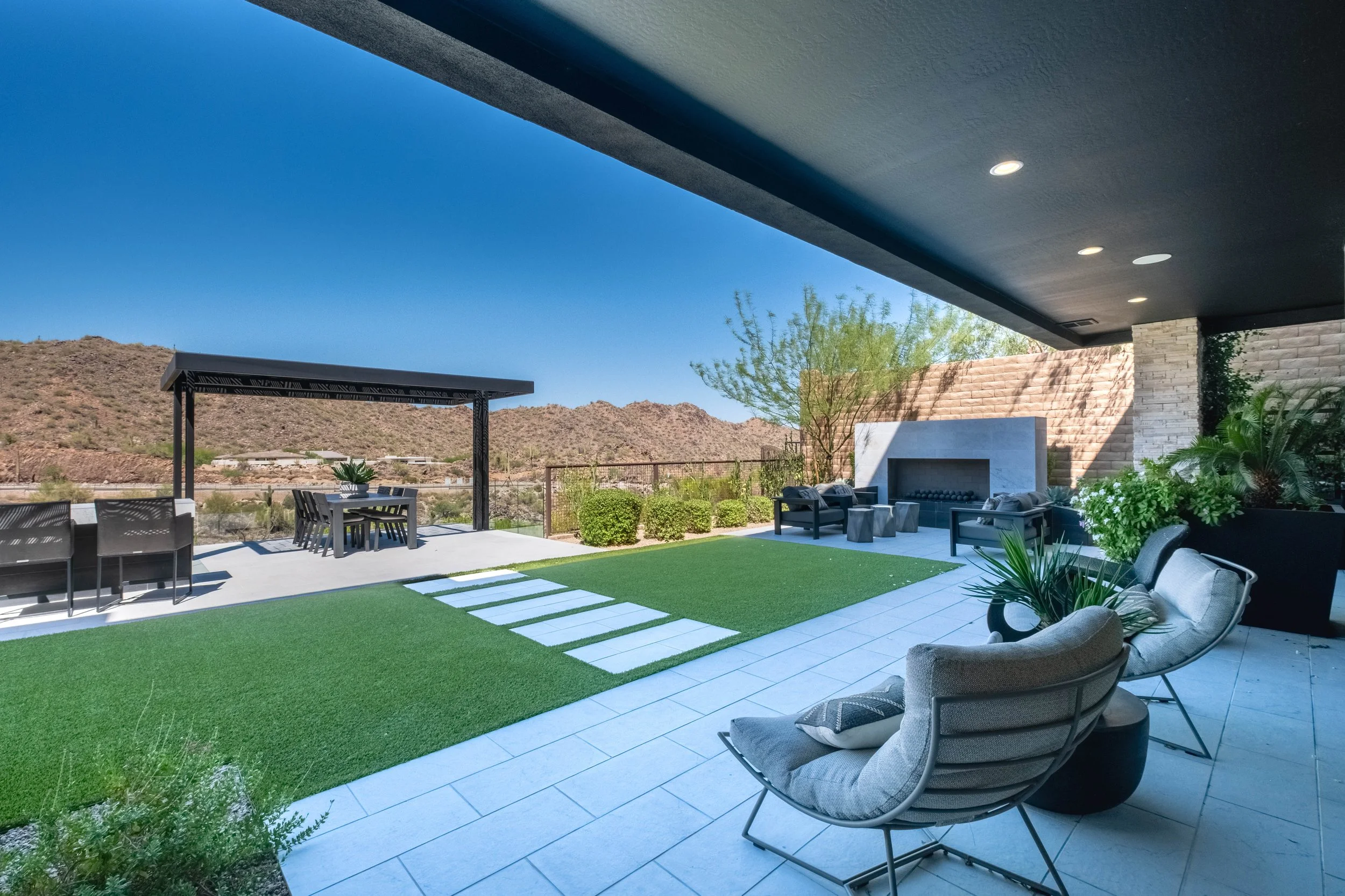 A modern outdoor patio with a covered ceiling, outdoor furniture including lounge chairs, a dining table with chairs, a built-in fireplace, and lush greenery, with desert hills in the background under a clear blue sky.