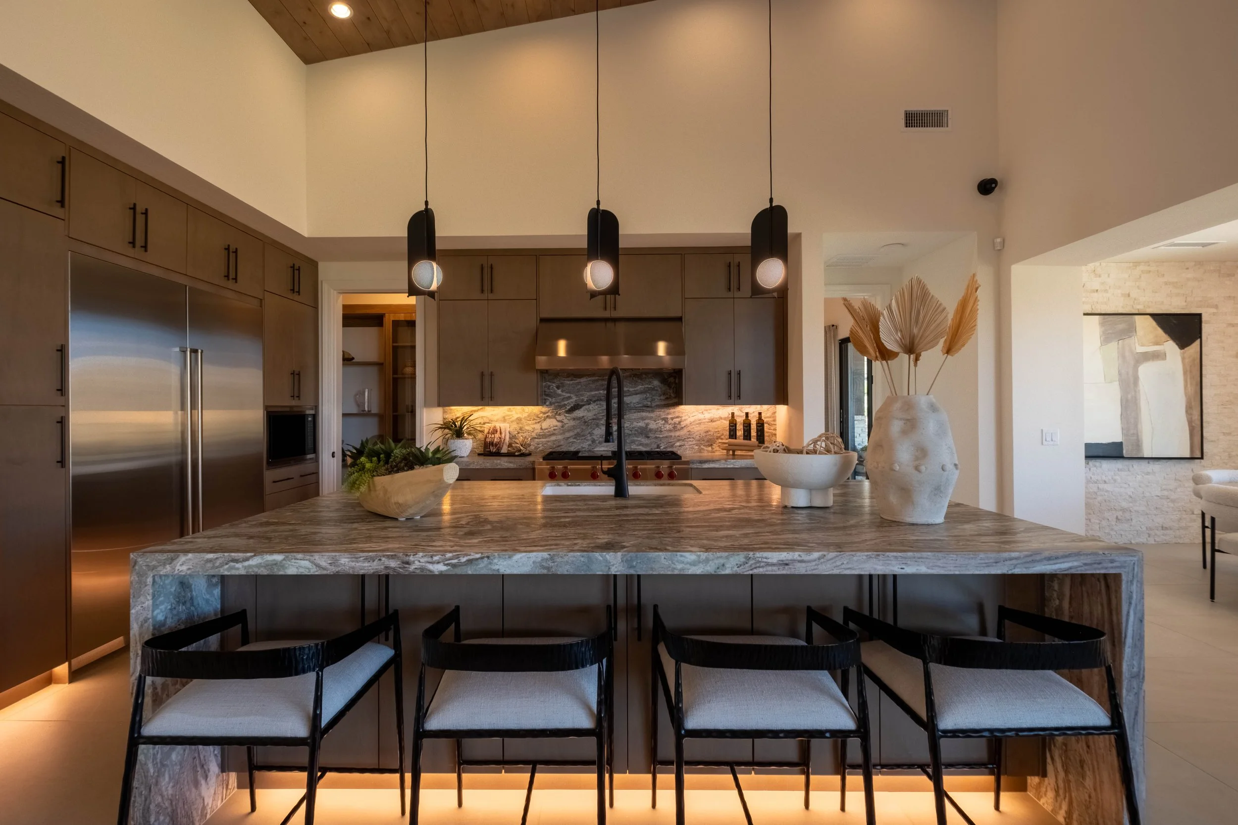 Modern kitchen with a marble island, black chairs, pendant lights, wooden cabinets, and decorative vases.