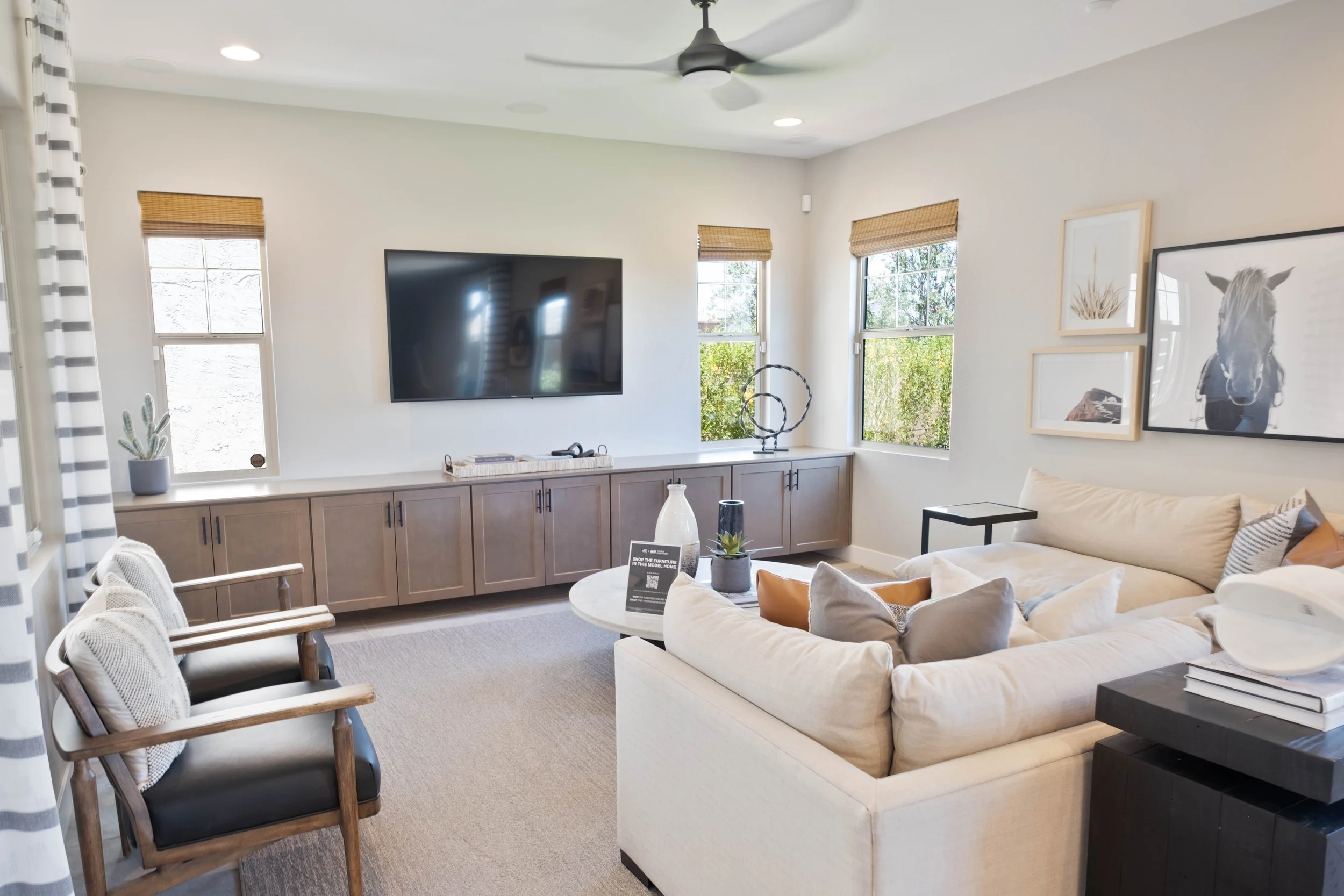 Modern living room with beige sofa, wooden chairs, a large flat-screen TV, and framed artwork on the wall. Natural light from multiple windows with bamboo blinds illuminates the space, which features a ceiling fan and a cabinet with decorative object