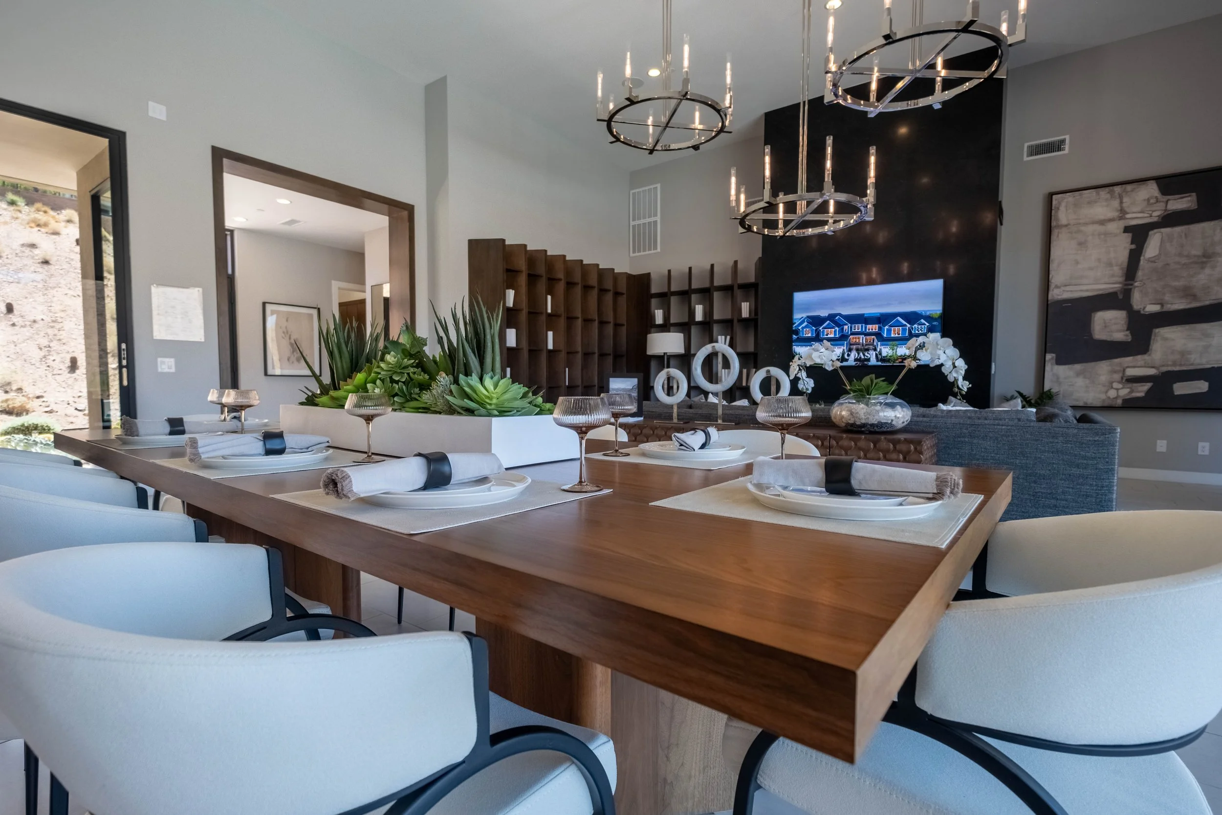 A modern dining room with a wooden table set with plates, glasses, and napkins. The room has white upholstered chairs, a large central succulent arrangement, and a wall mounted TV. There are pendant lights hanging from the ceiling, and decorative art