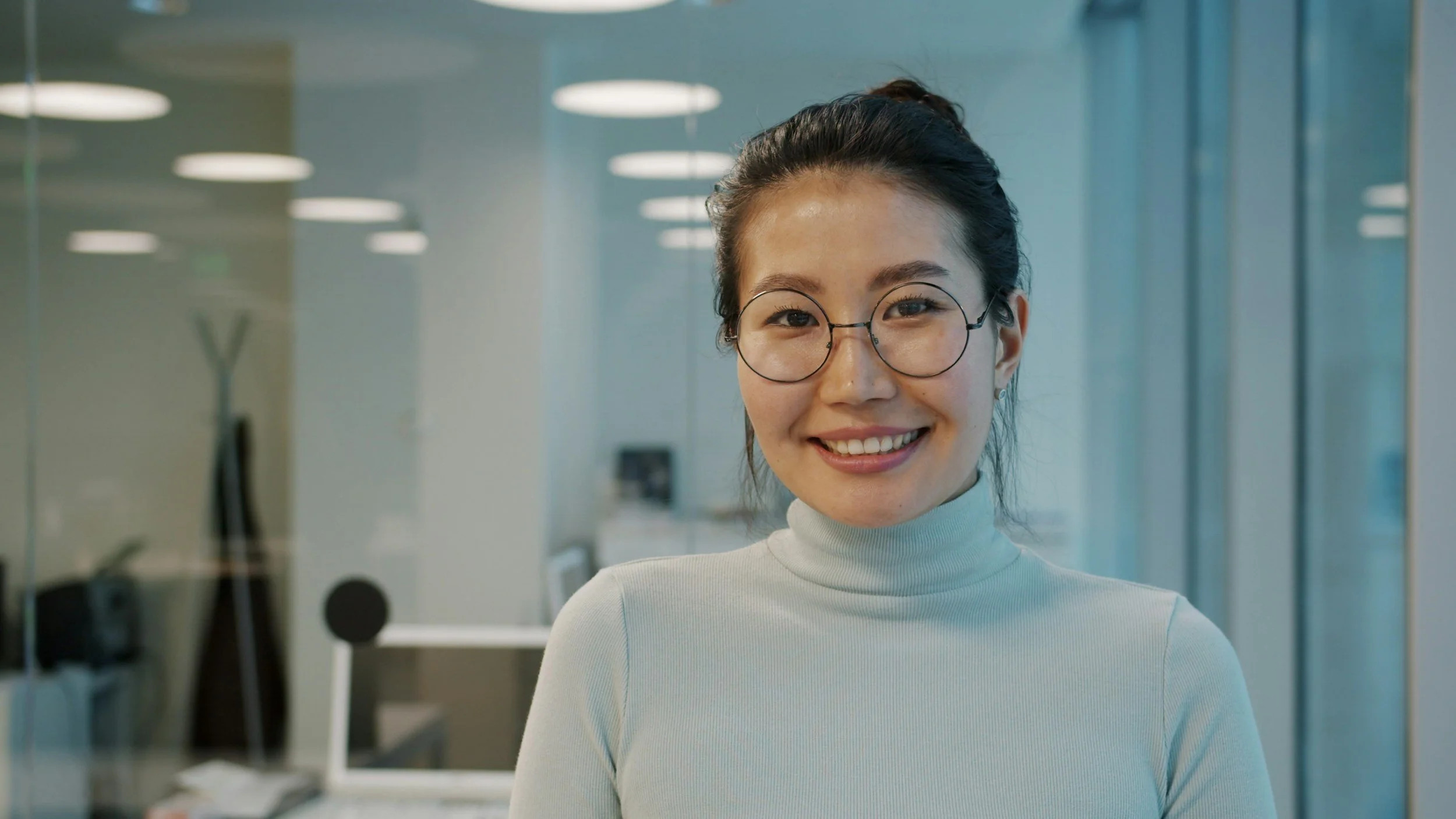 A young woman with dark hair tied back, wearing round glasses and a white high-neck top, smiling in an office with glass walls and modern lighting.