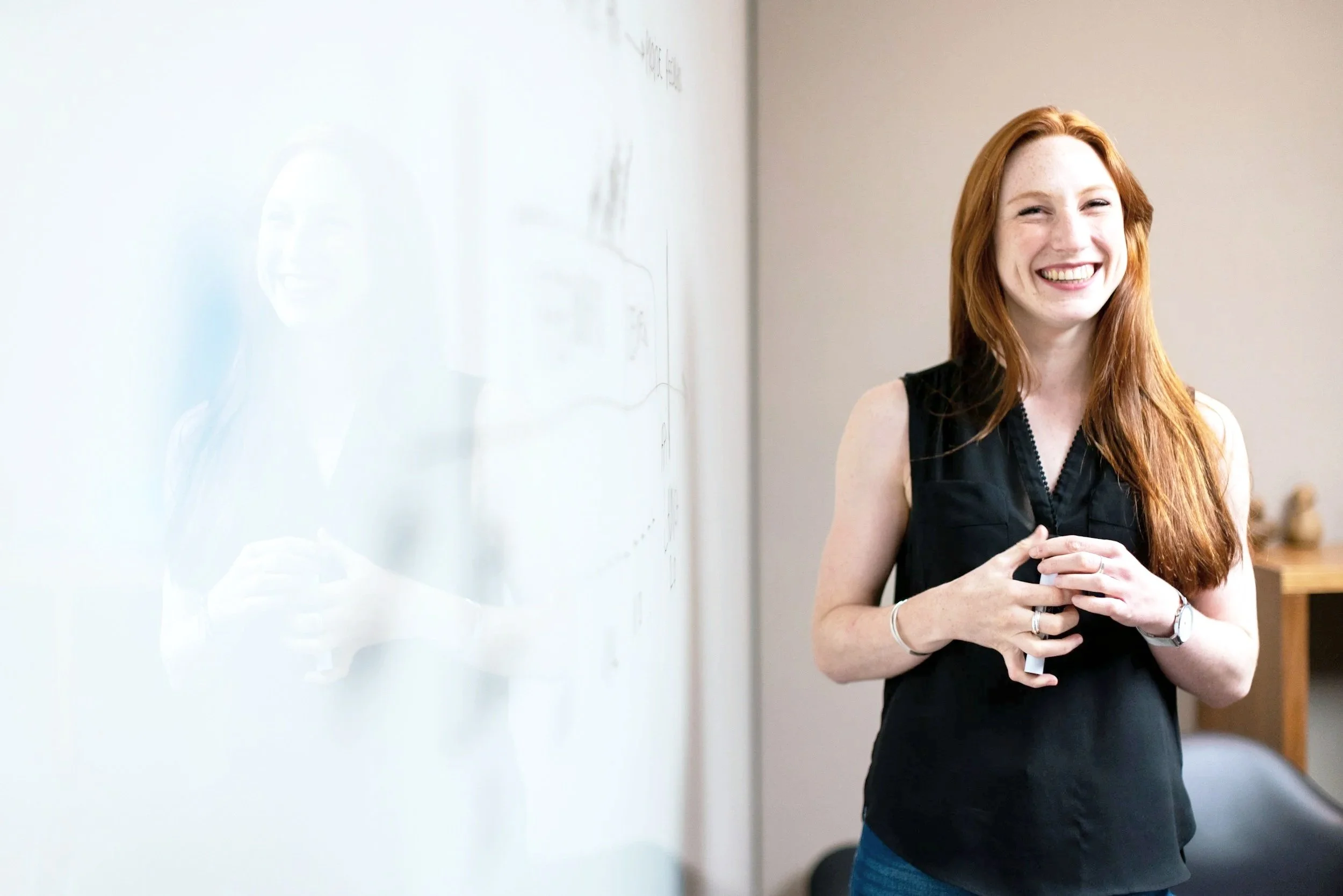 A woman with long red hair smiling and standing next to a whiteboard.