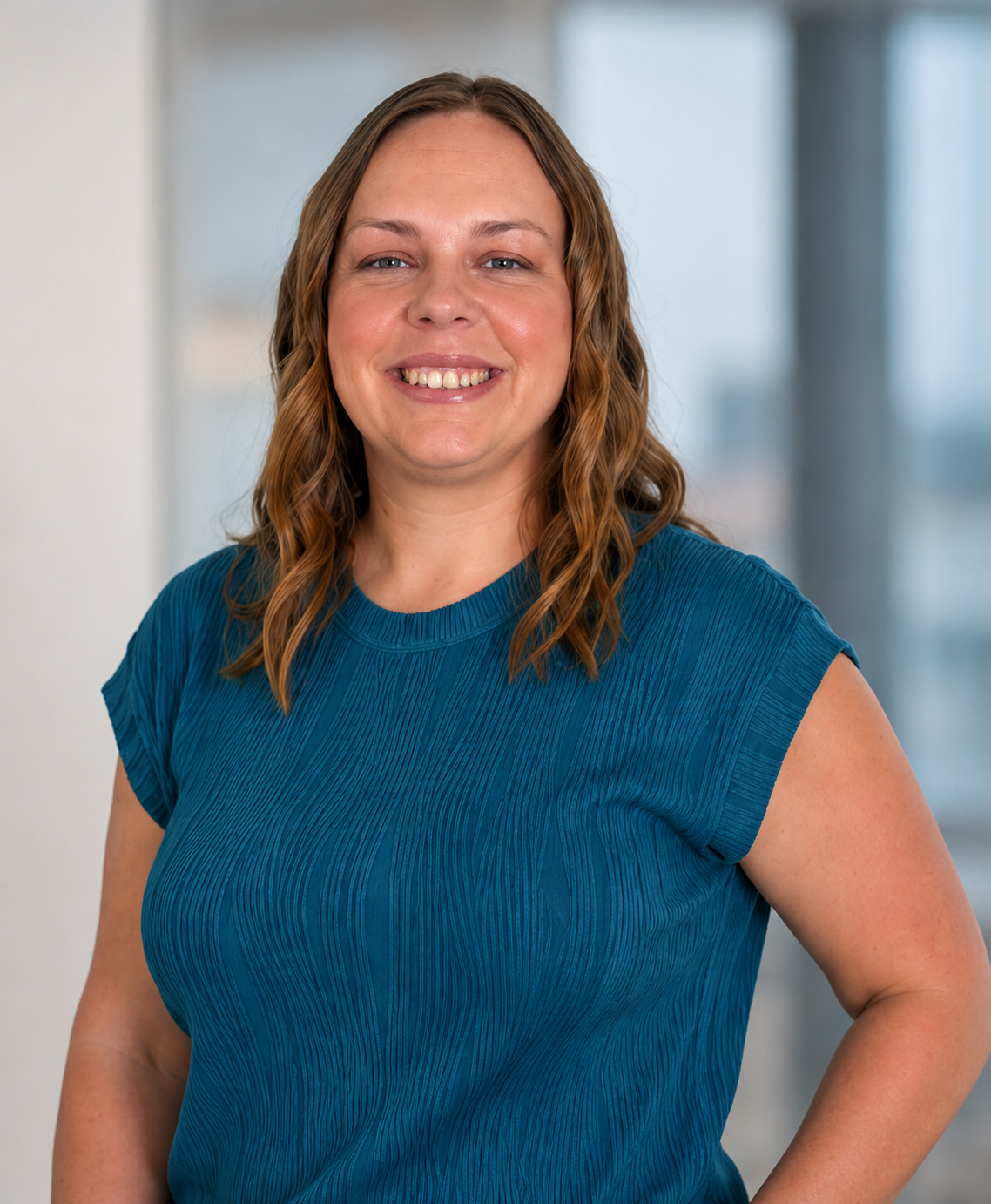 Becca Bradd. A smiling woman with shoulder-length wavy blonde brown hair, wearing a blue textured short-sleeve top, standing in front of a blurred indoor background with large windows.
