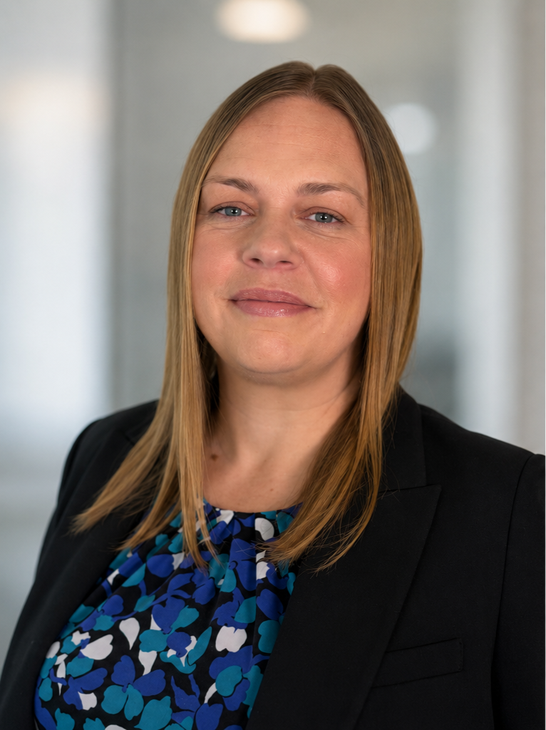 Becca Bradd. A professional woman with straight, blonde brownhair wearing a black blazer and blue patterned blouse, standing in an office setting with a neutral background.