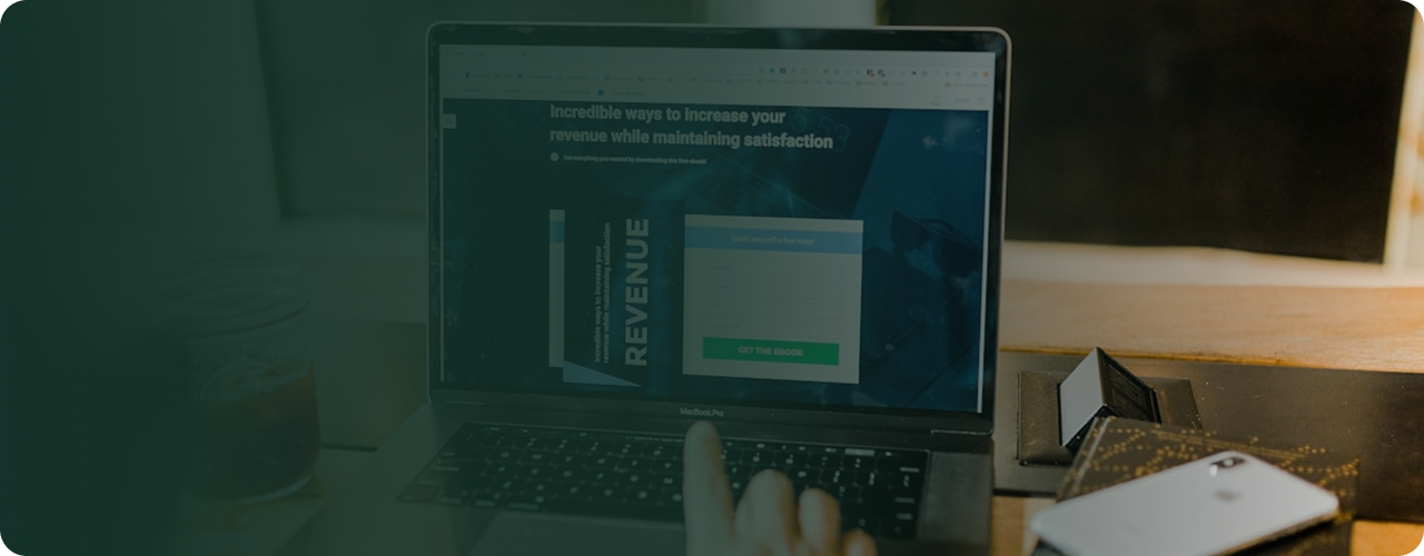 A person using a MacBook Pro laptop with a website about increasing revenue displayed on the screen, in a dimly lit workspace with a jar, smartphone, and a small display stand nearby.