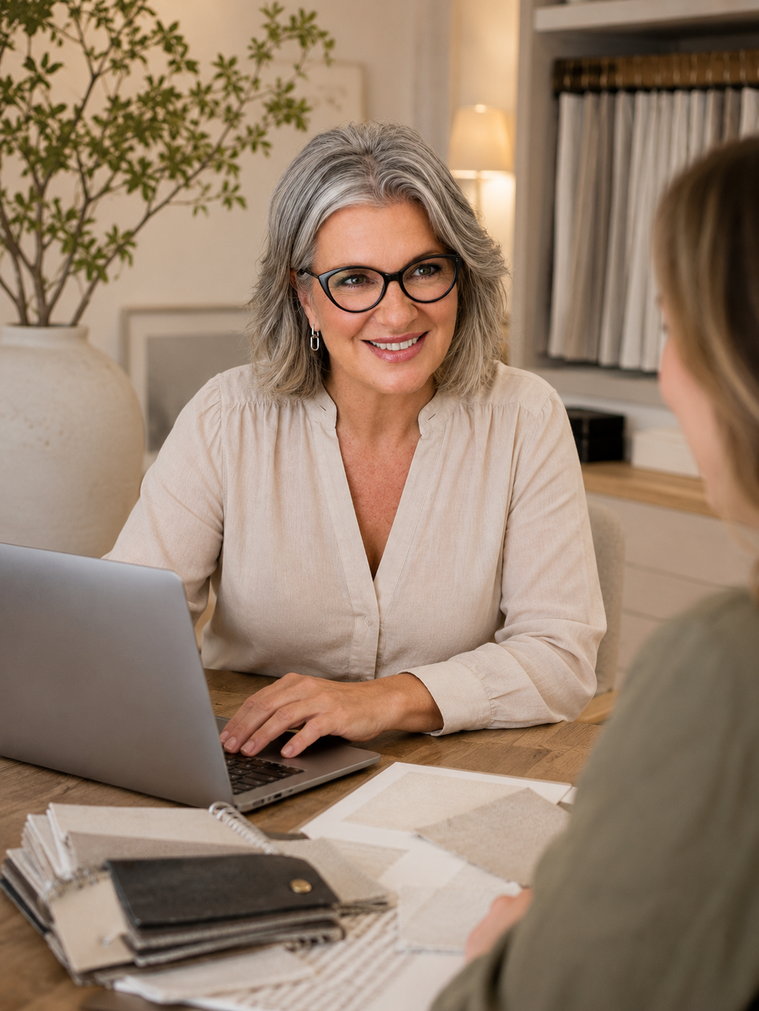 A woman with gray hair, glasses, and earrings smiling while talking to another person in an office setting. The woman is sitting at a desk with a laptop, color swatches, and a notebook. In the background, there is a large potted plant, books, and a lamp.
