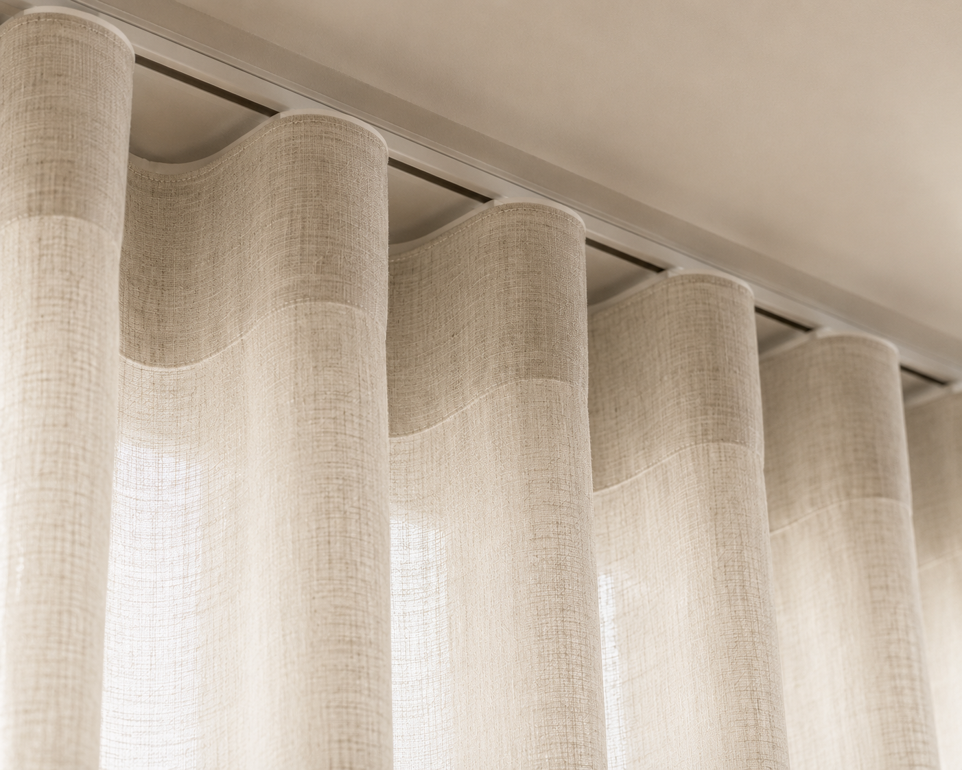 Light beige curtains hanging in a S-wave pleat formation, from a metal curtain track that is top-fit mounted to the ceiling.