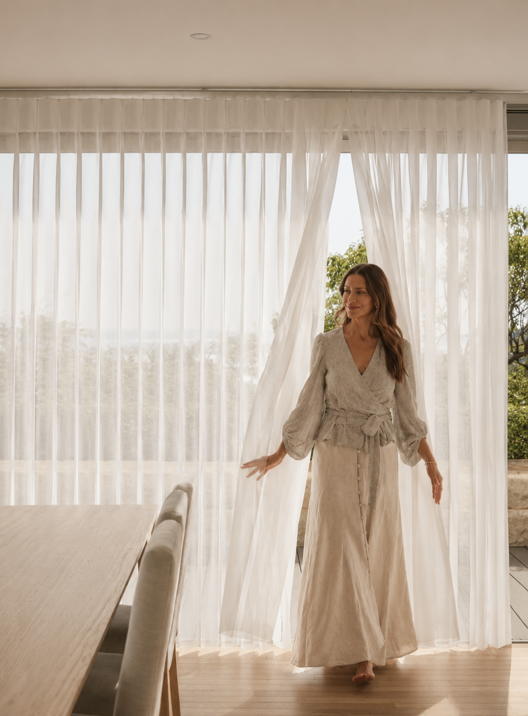 Woman in beige dress standing near sheer curtains in a bright room with outdoor greenery visible outside.