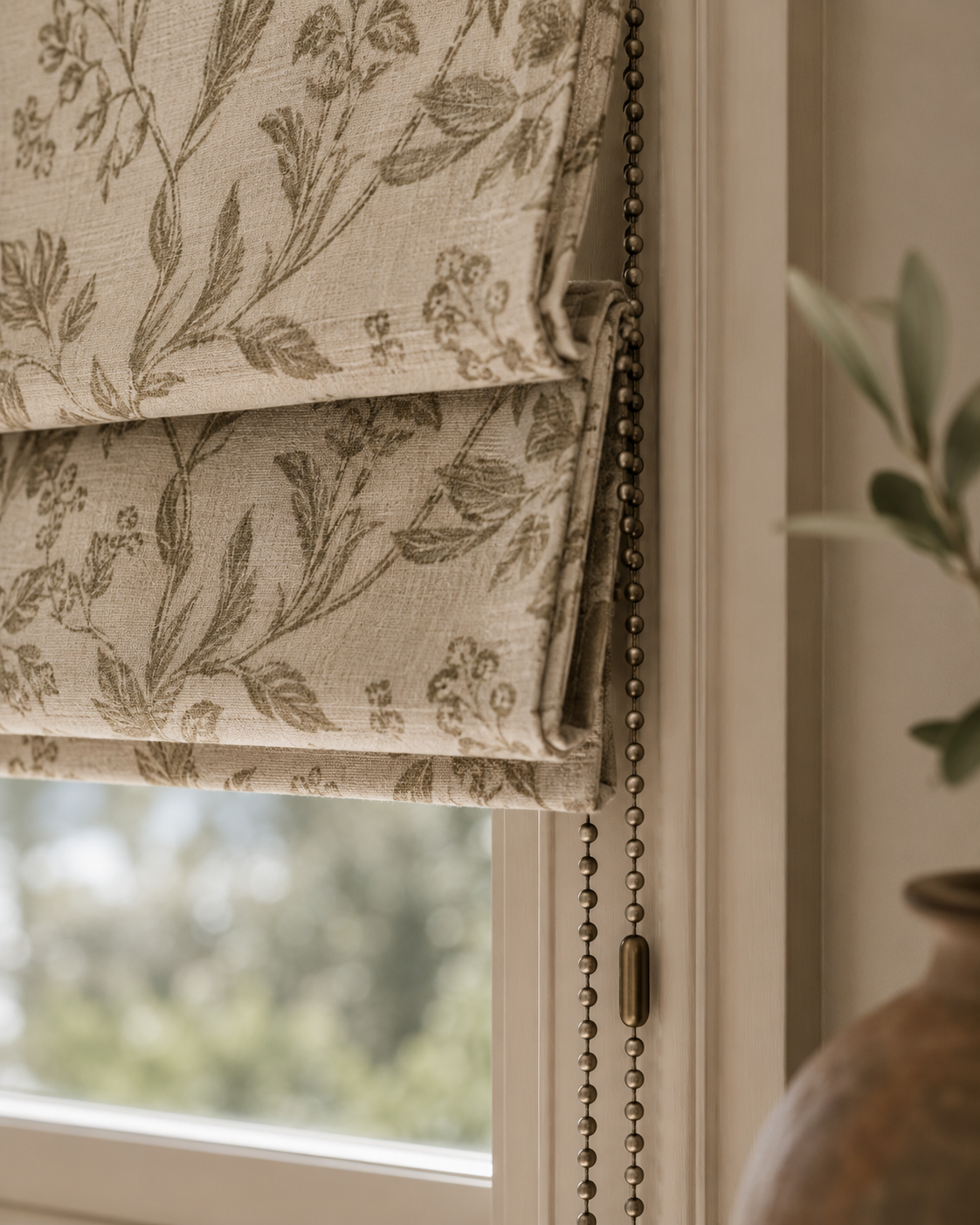 Close-up of a window with beige floral patterned Roman shade and a bead chain pull control, with a blurred outdoor view and a potted plant in the background.