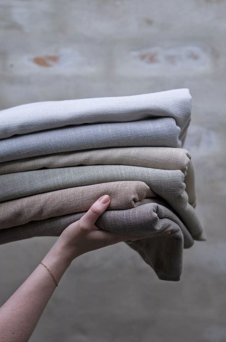 A person holding a stack of folded beige, light gray, and off-white fabric or clothing against a textured gray brick wall background.