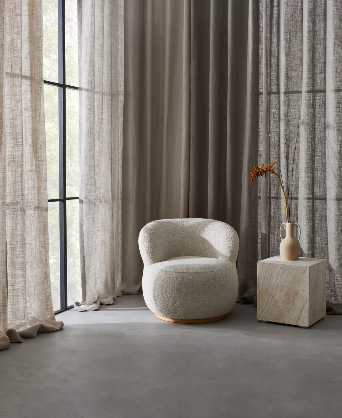 A cozy room with sheer and solid curtains, a plush white armchair, and a wooden side table with a cream vase holding dried orange flowers.