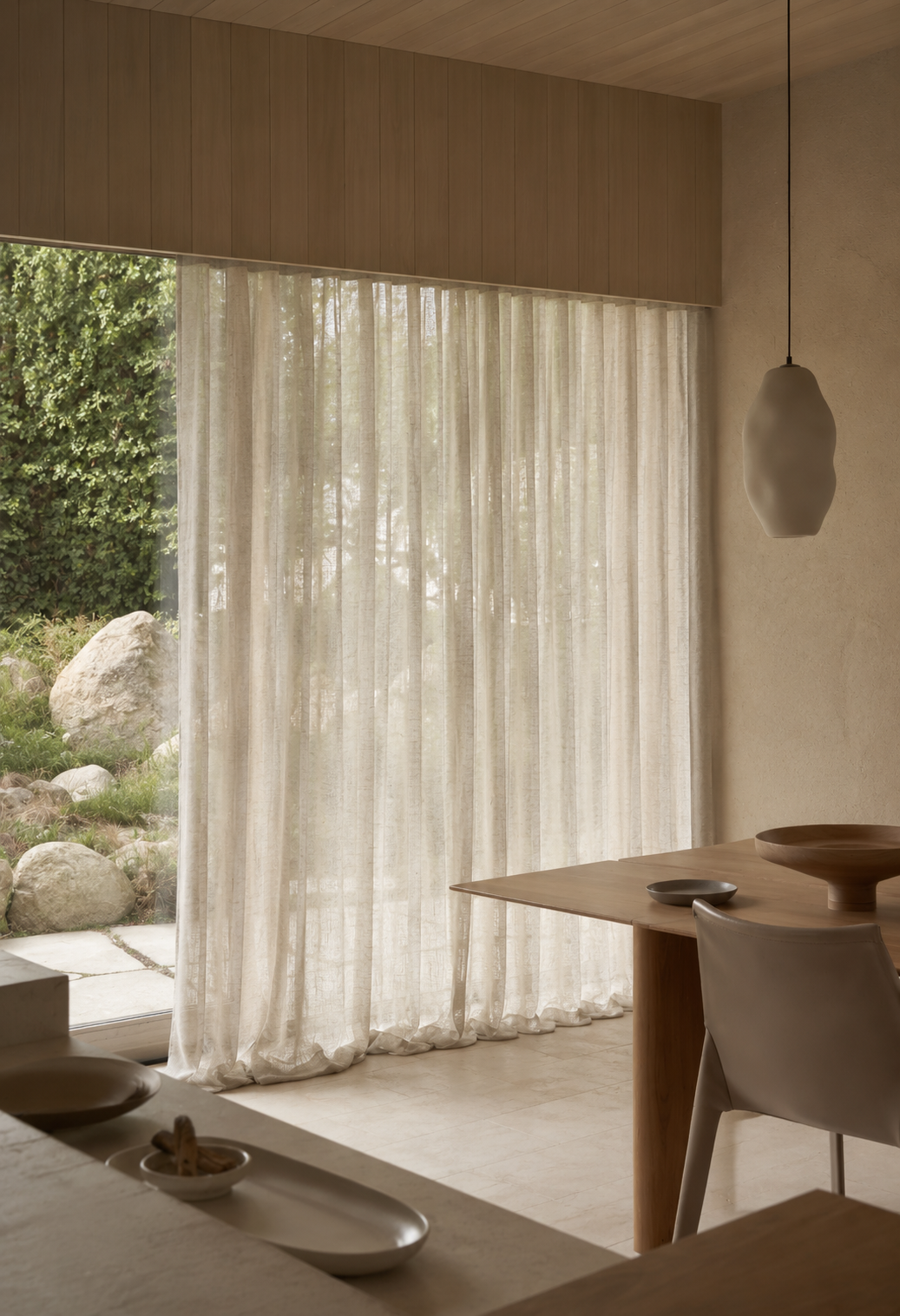 A room with large window covered by beige sheer curtains, showing a garden with rocks and greenery outside. The room features a wooden table with a white chair, and a hanging pendant light.